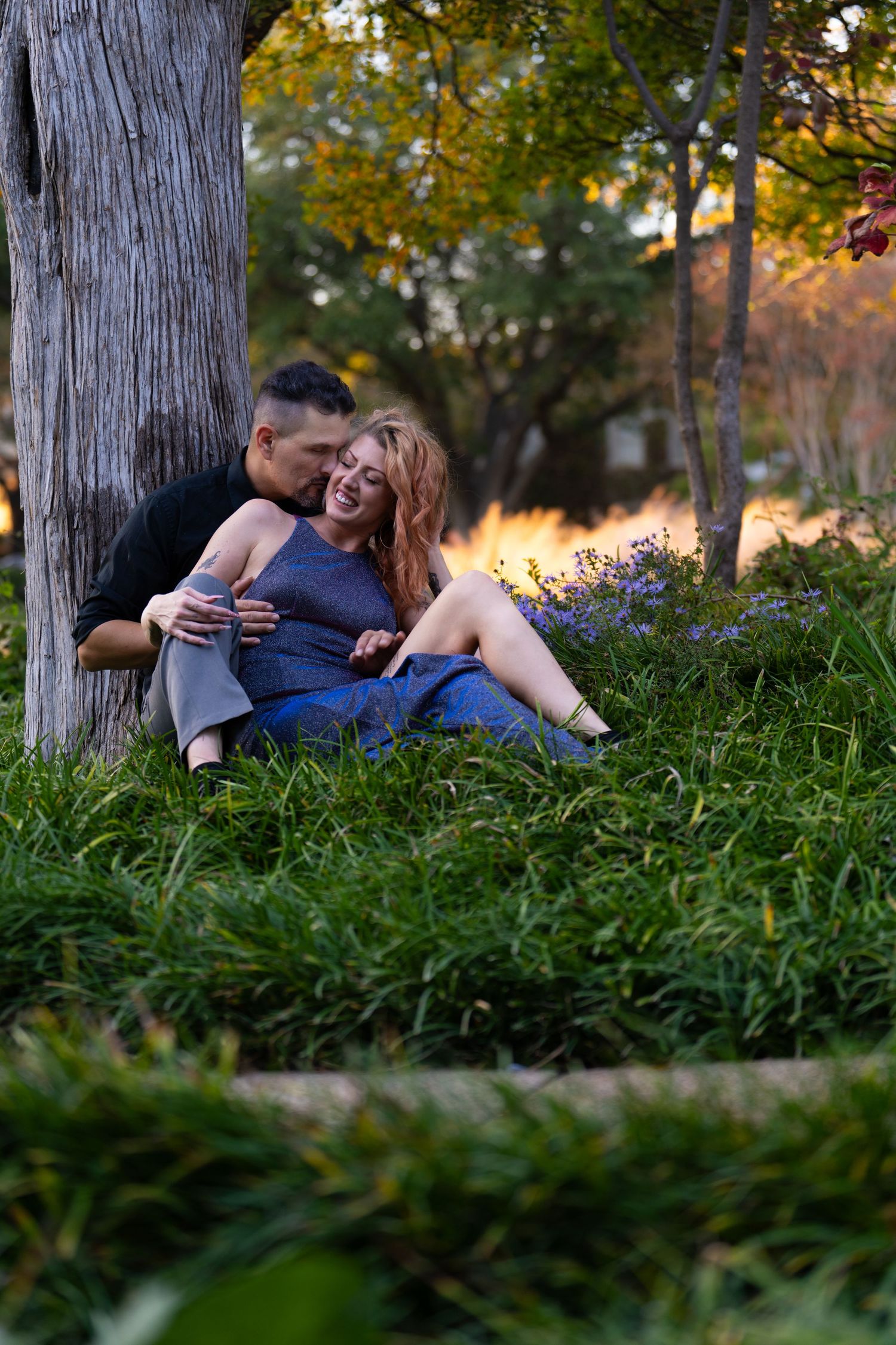 Intimate moment captured between couple sitting on grass beneath tree at sunset.
