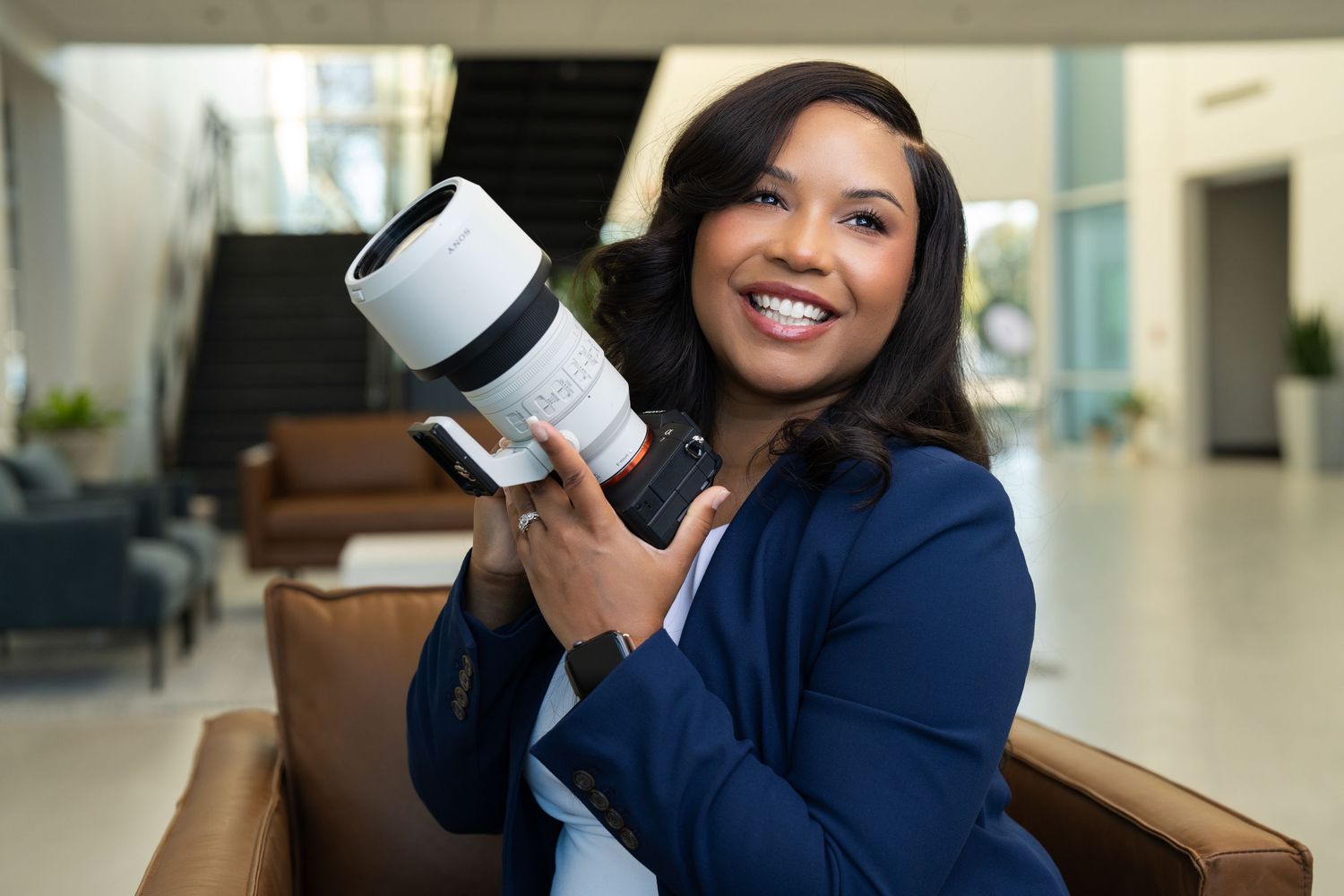 Two smiling coworkers in business attire hold coffee cups while having a conversation in a bright office hallway.