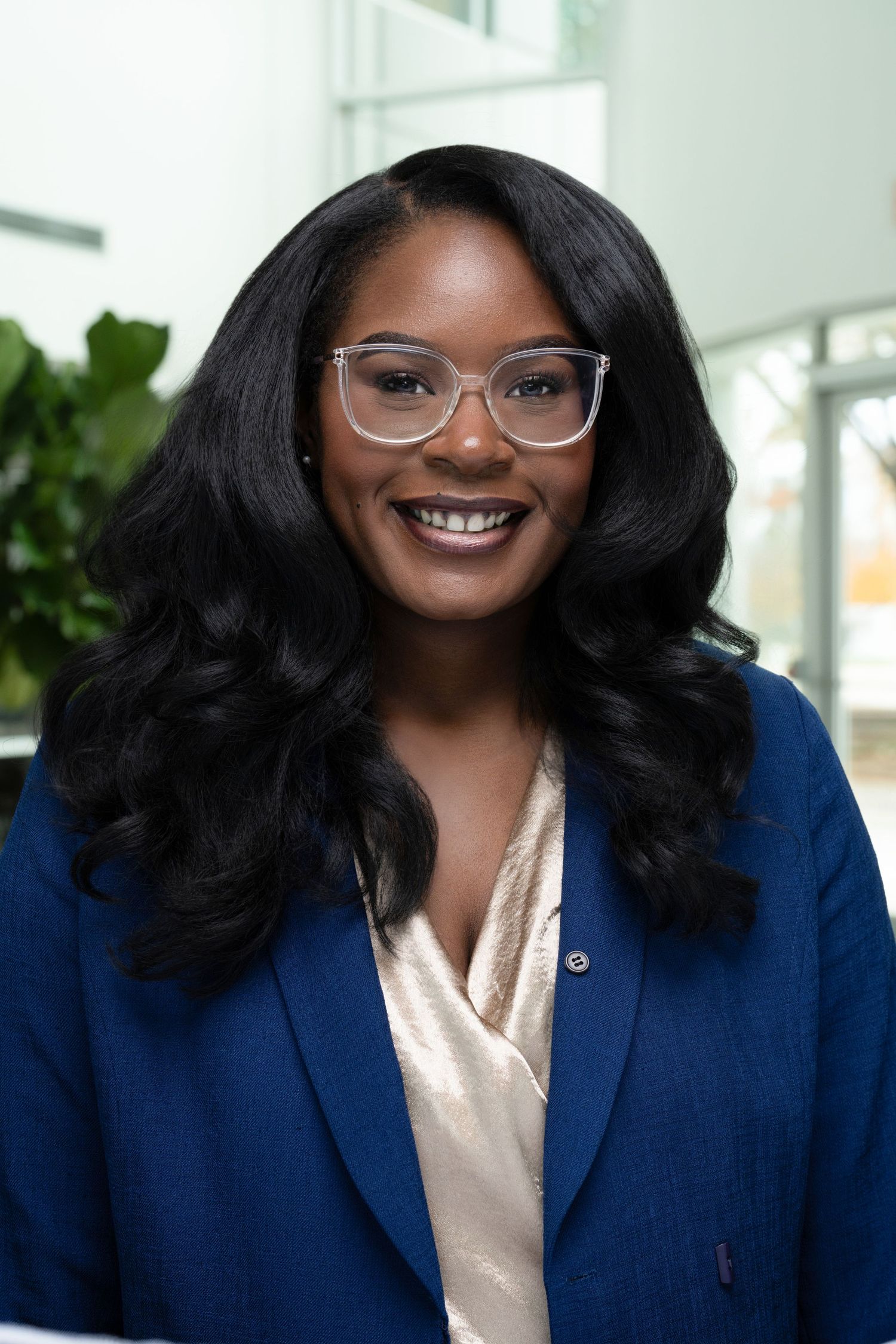 Professional portrait showing someone in a navy blazer and white blouse wearing glasses against a light office background.