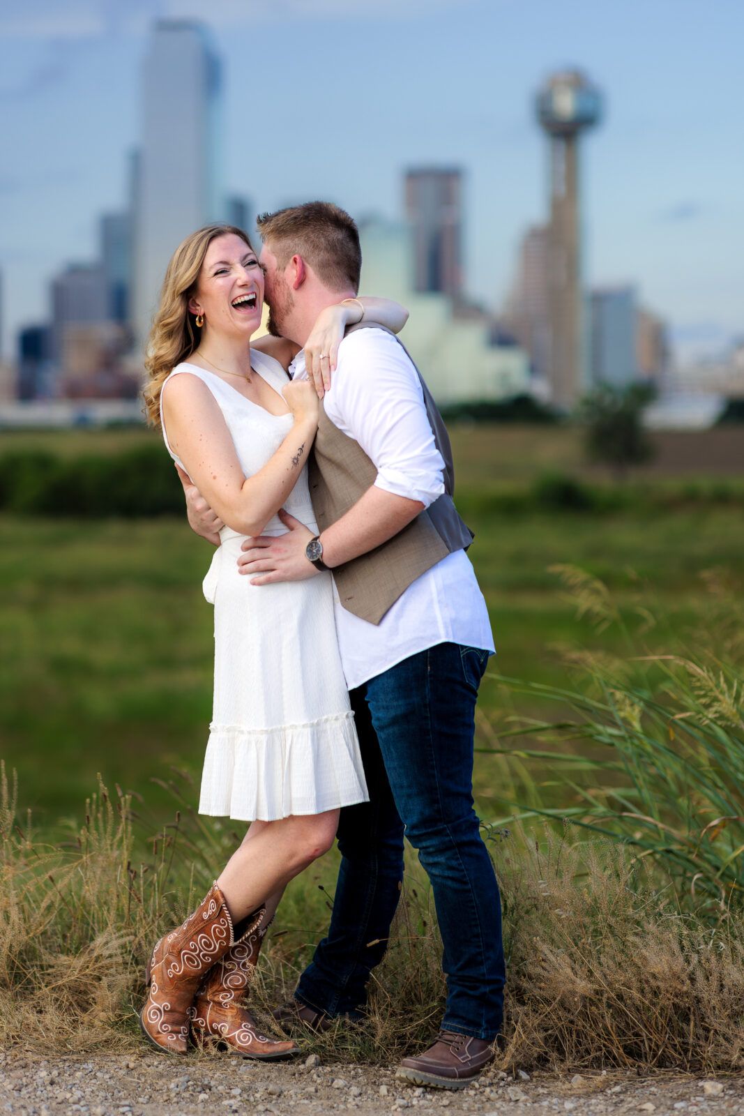 Couple sharing an intimate moment near a city skyline with boots and grass in the foreground.