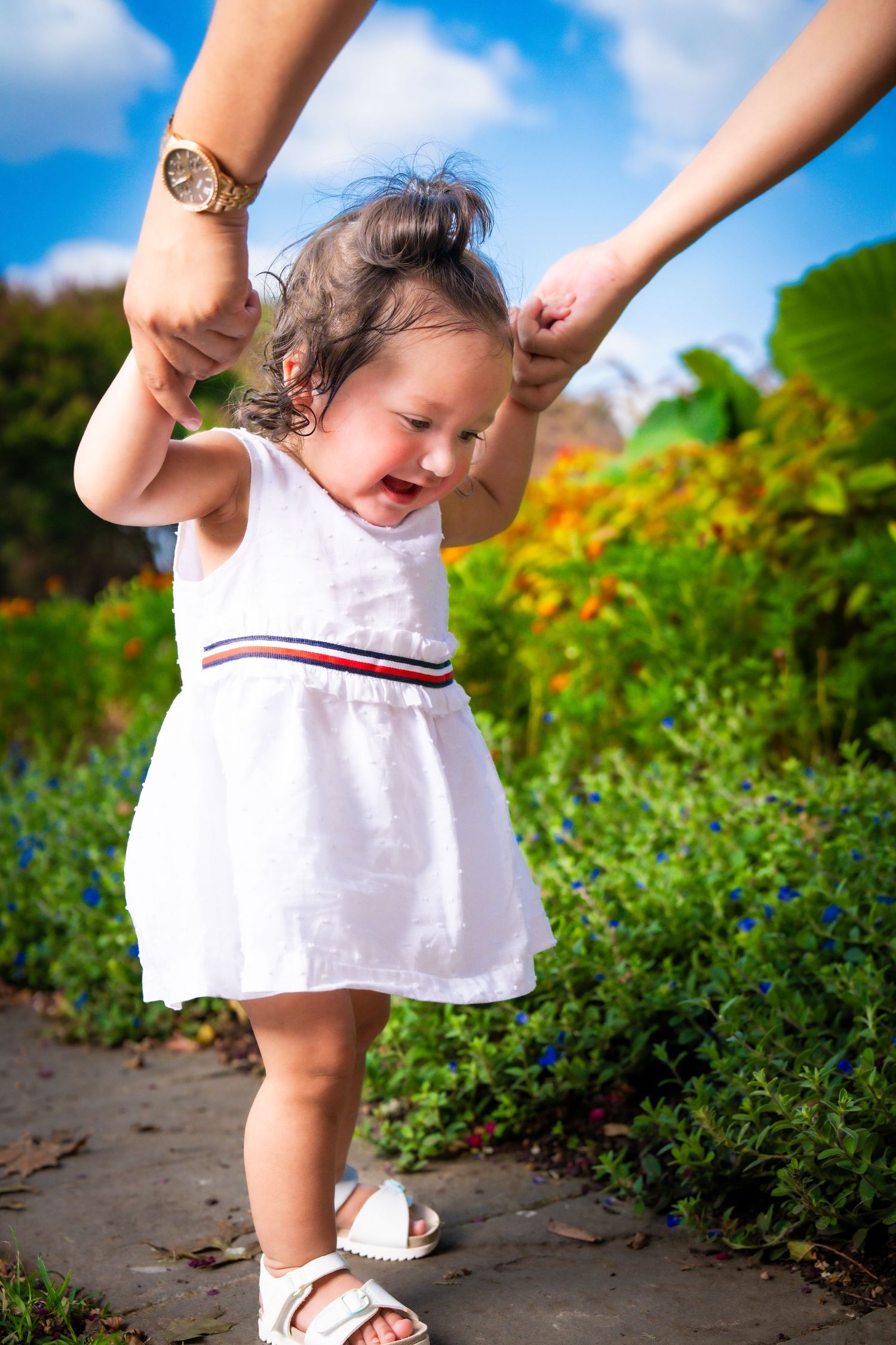 Toddler in white dress holding hands and walking along garden path.
