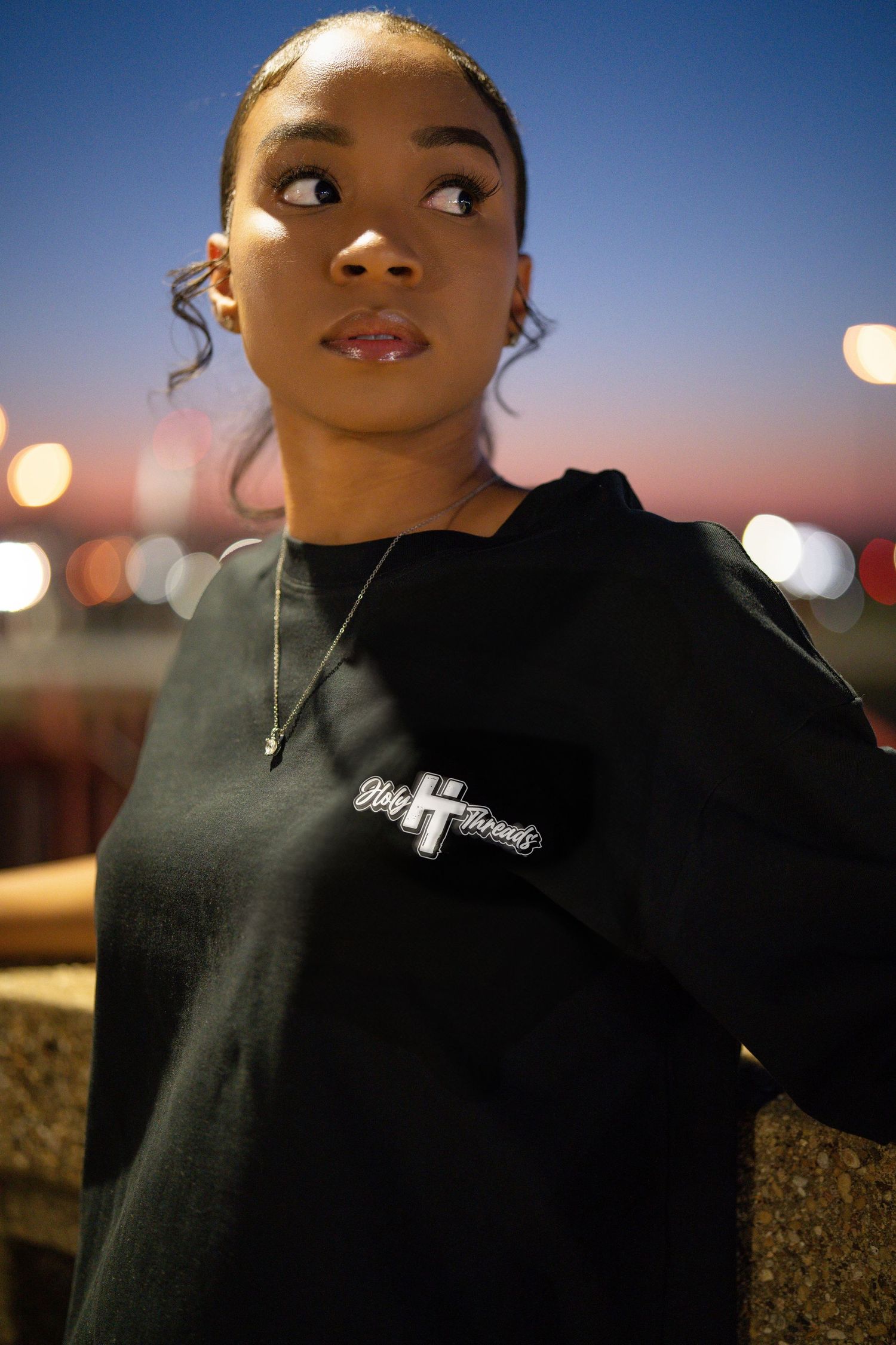 Person wearing black Holy Threads hoodie against twilight sky with bokeh lights.