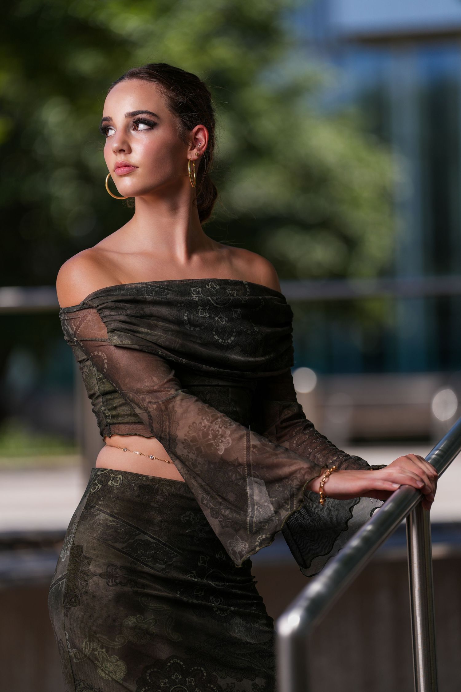 A dramatic fashion portrait featuring a sheer black off-shoulder dress against an urban background.