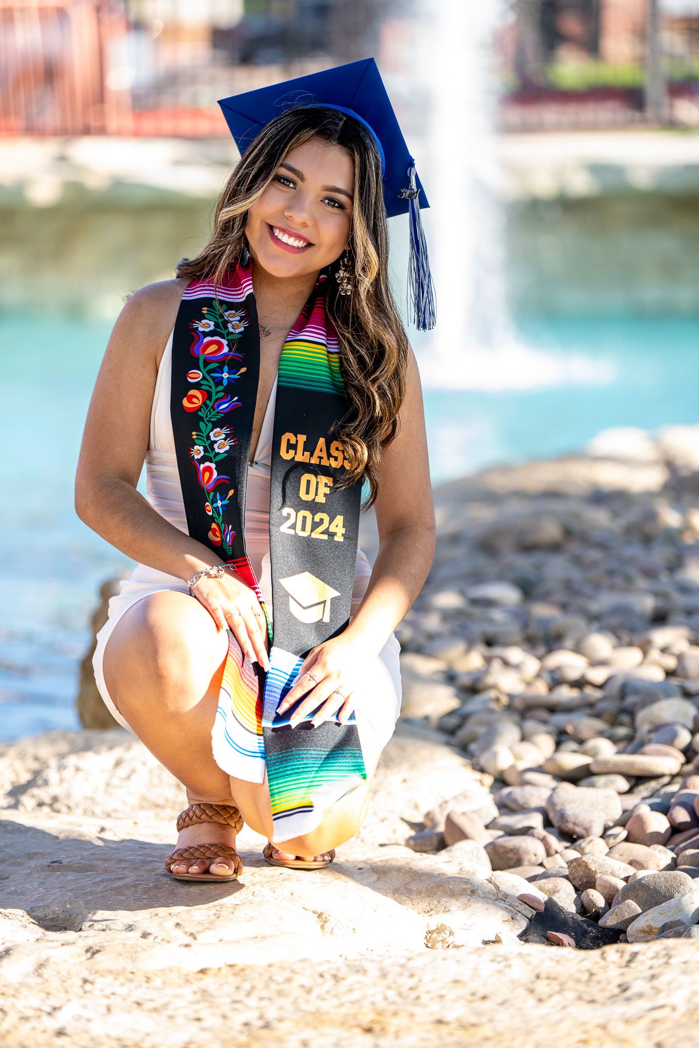Graduate in blue cap and colorful sash poses by water feature in white dress.