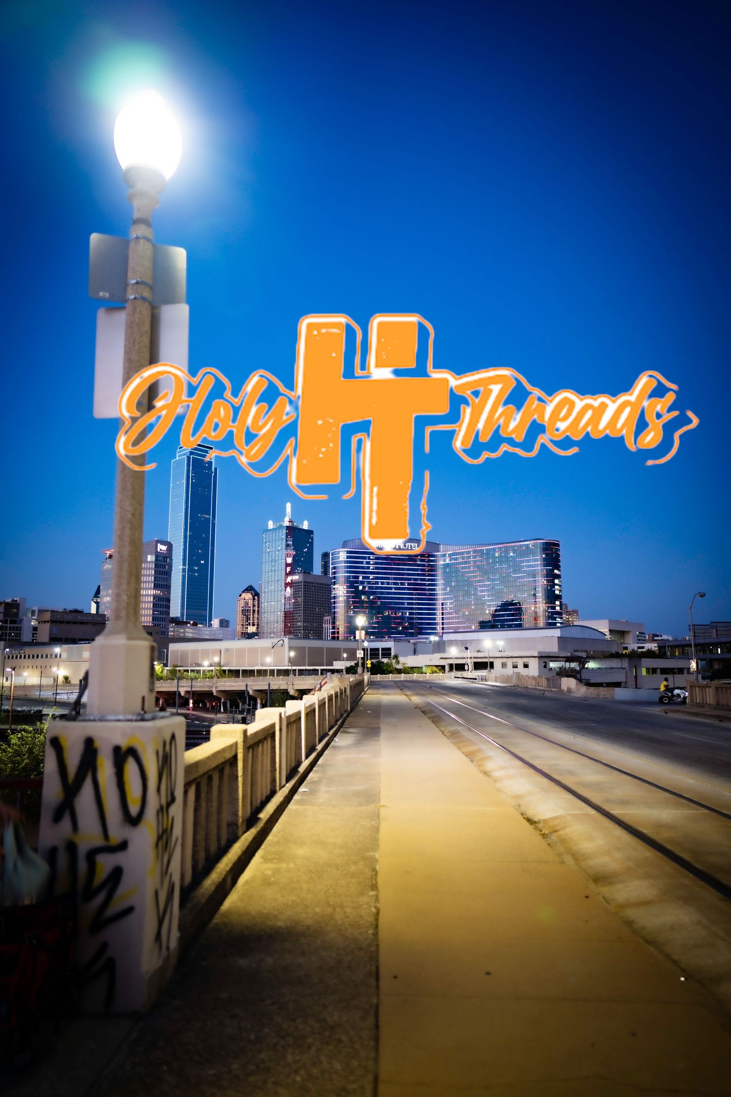Holy Threads illuminated sign over city skyline at dusk with street lamp in foreground.