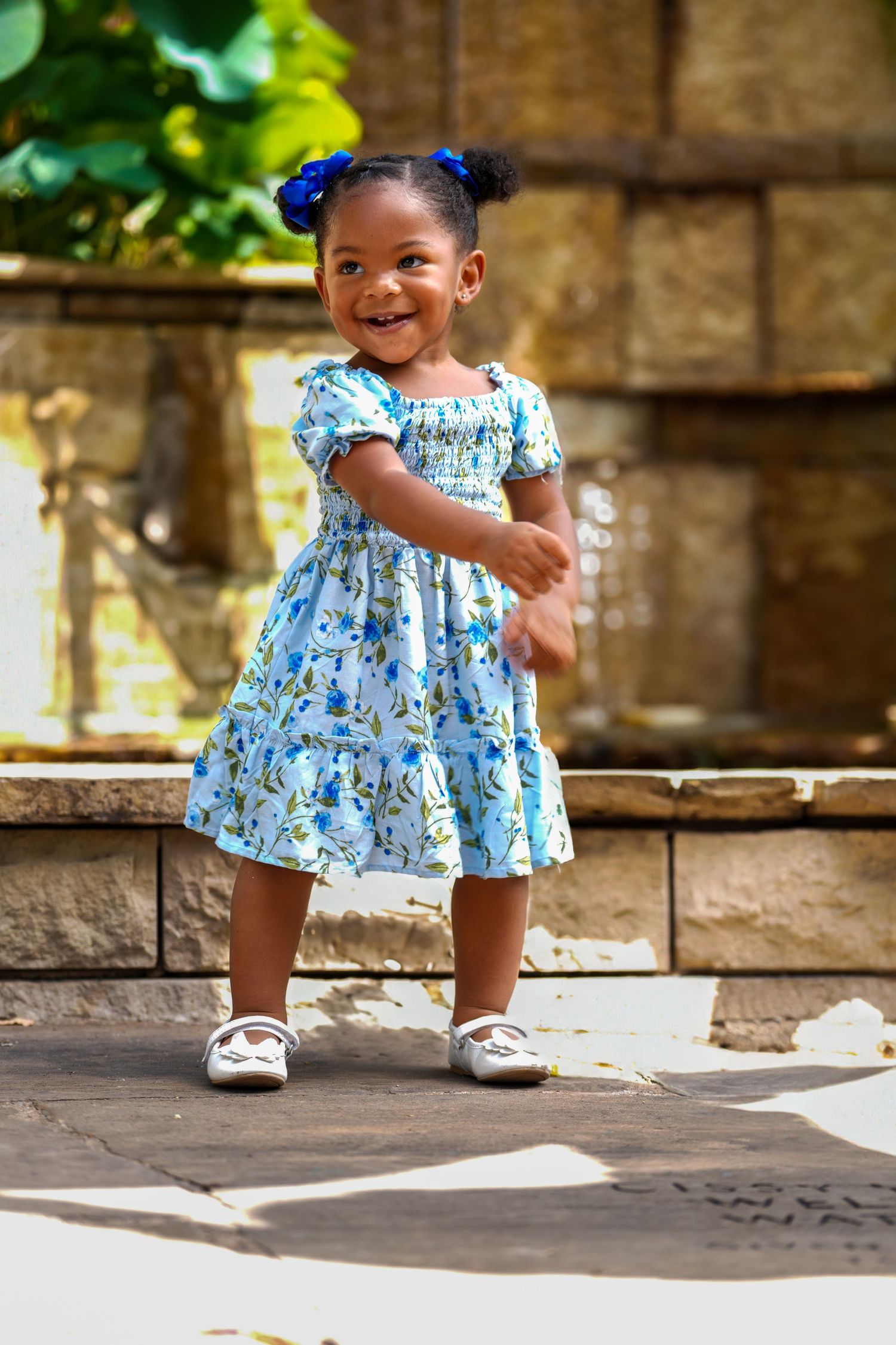 Small child wearing blue floral dress and white shoes poses against stone wall backdrop.