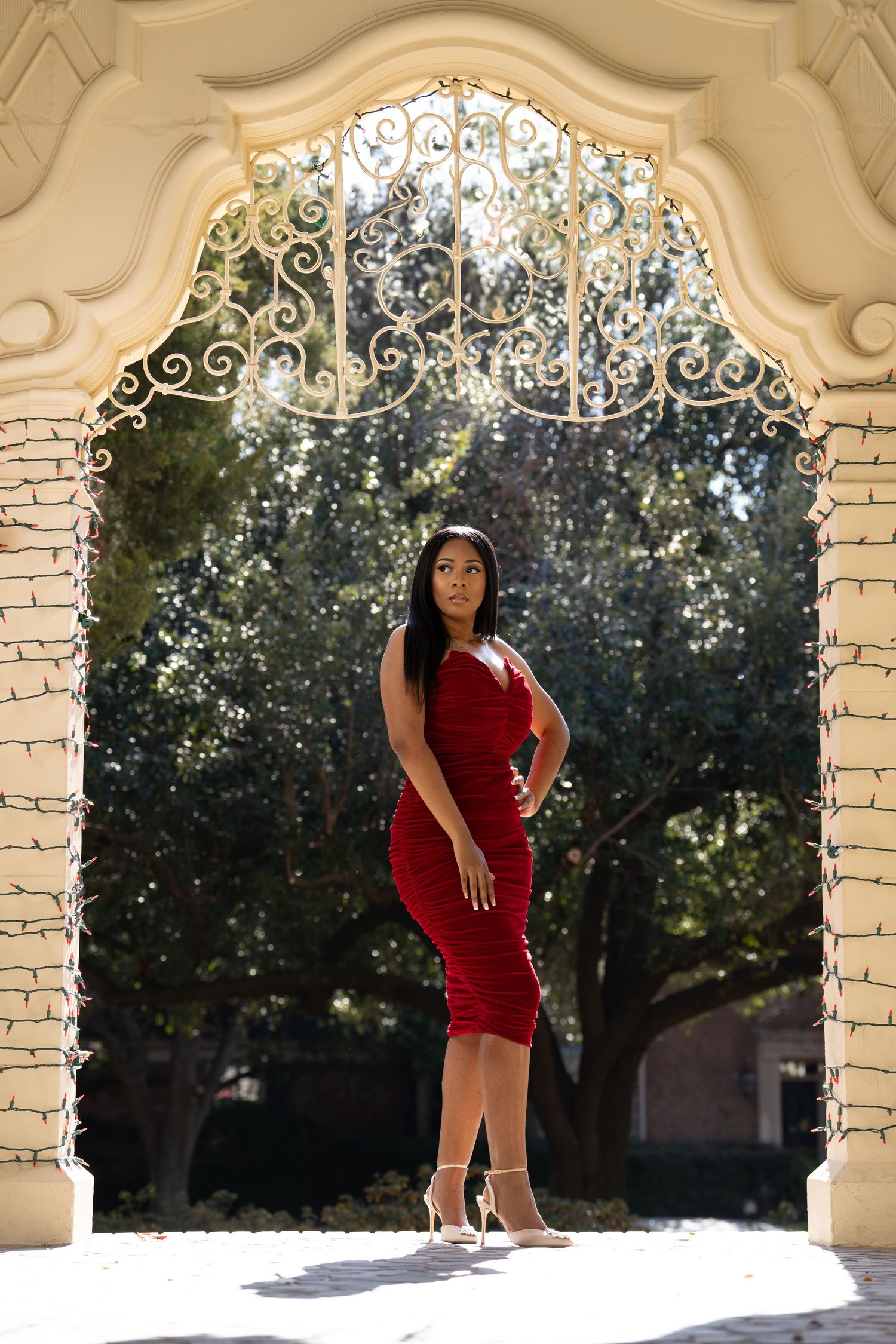 An elegant figure poses in a form-fitting red dress under an ornate archway with decorative ironwork and natural lighting.