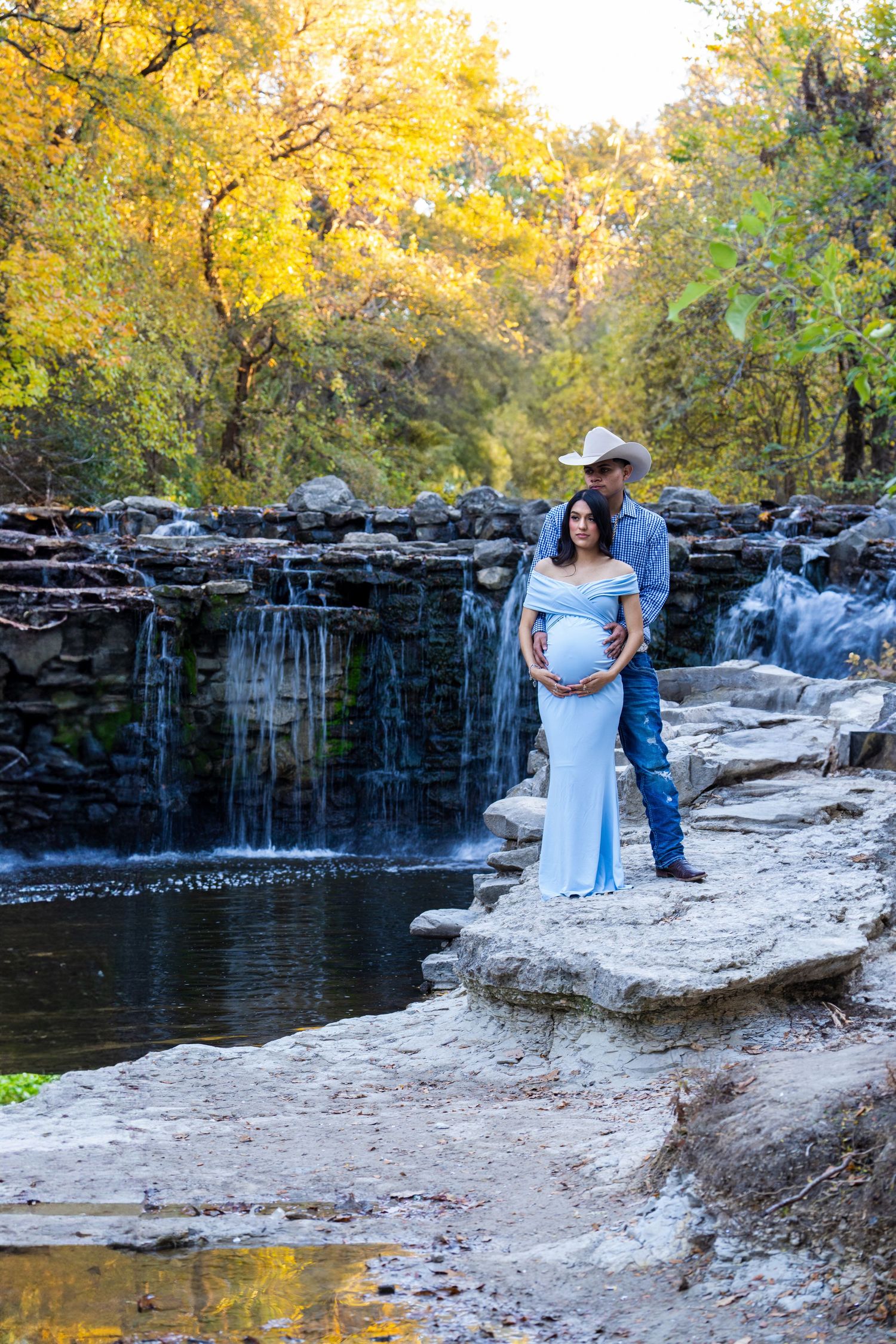 Couple stands together on rocky outcrop by cascading waterfall surrounded by autumn foliage in golden afternoon light.