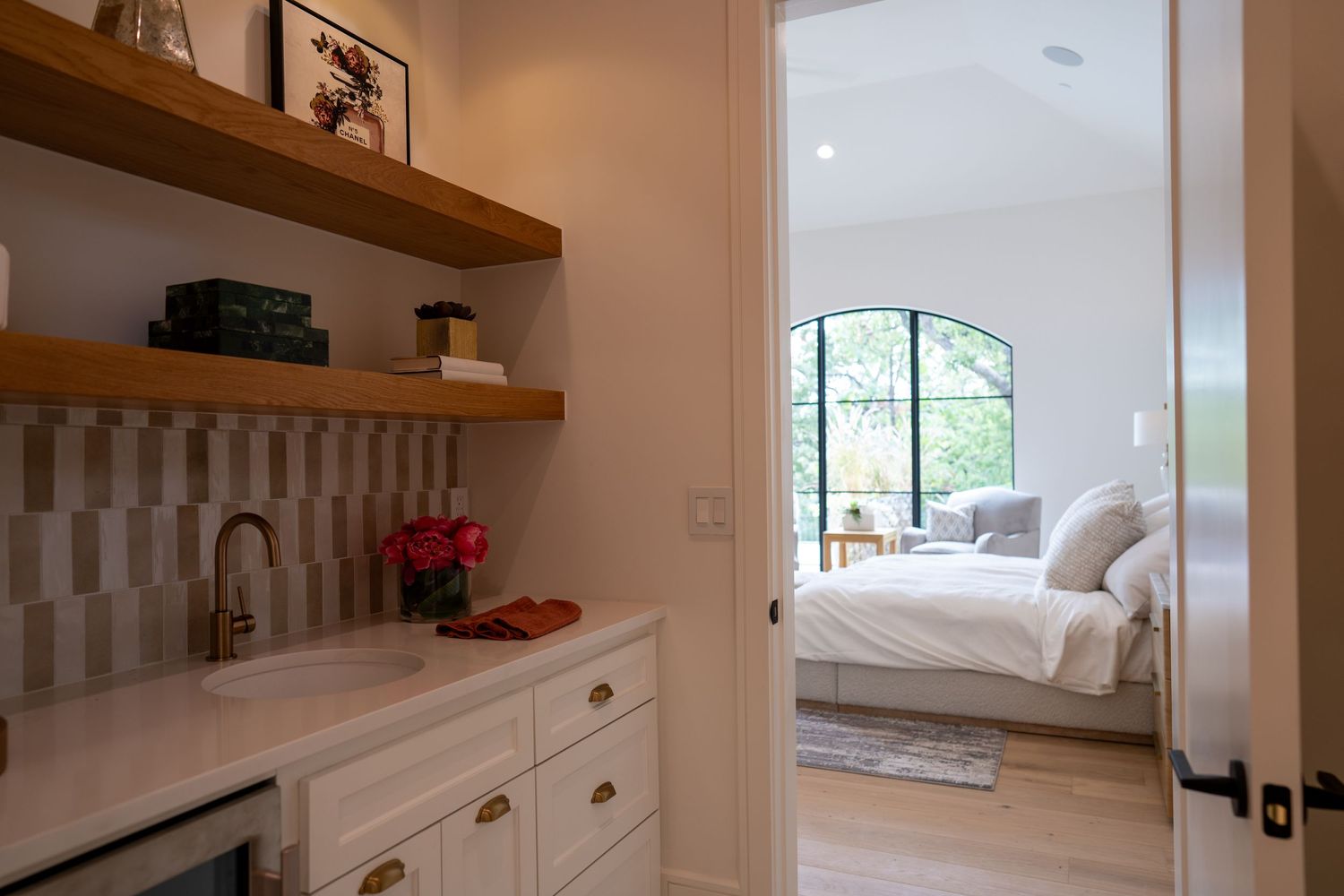 Cozy bedroom suite with attached kitchenette featuring white cabinets and wooden floating shelves.