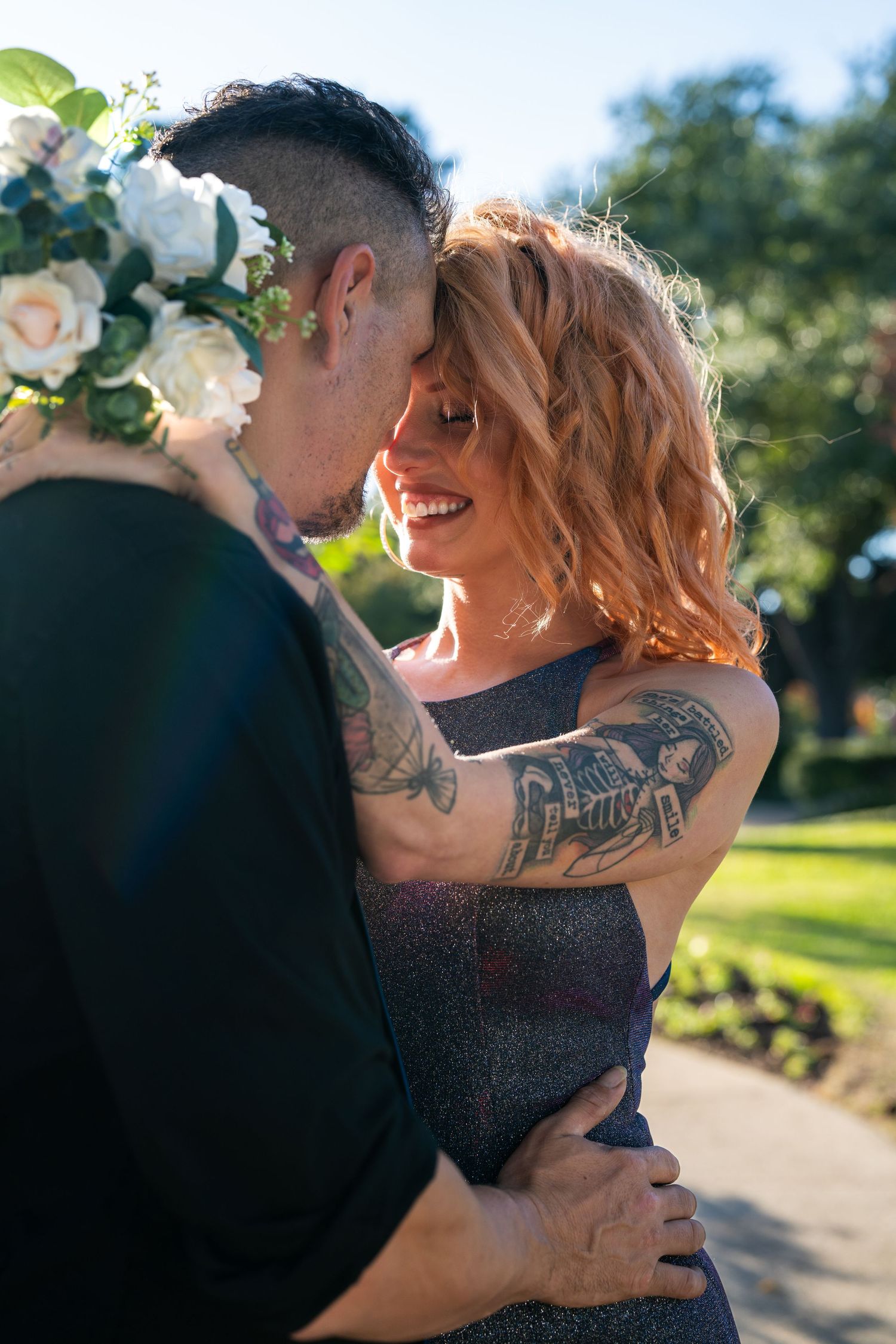A romantic couple embraces outdoors while holding a bouquet of white flowers.