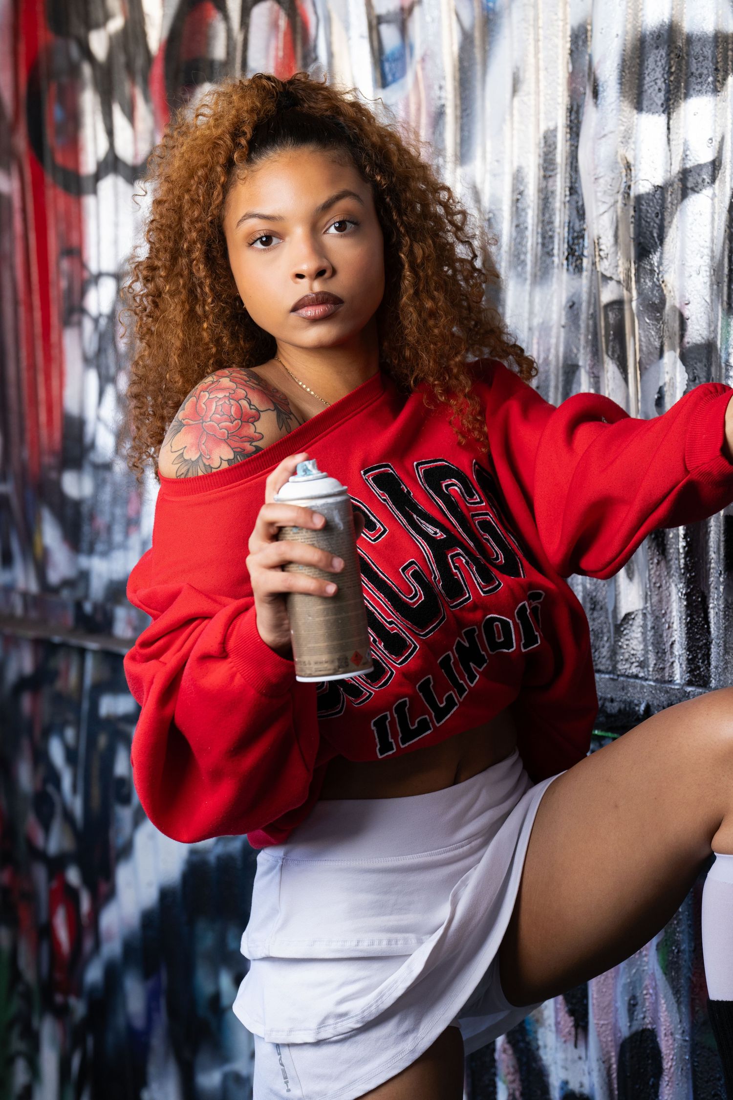 Young woman in red sweatshirt holding spray paint can poses against graffiti wall in urban photoshoot.