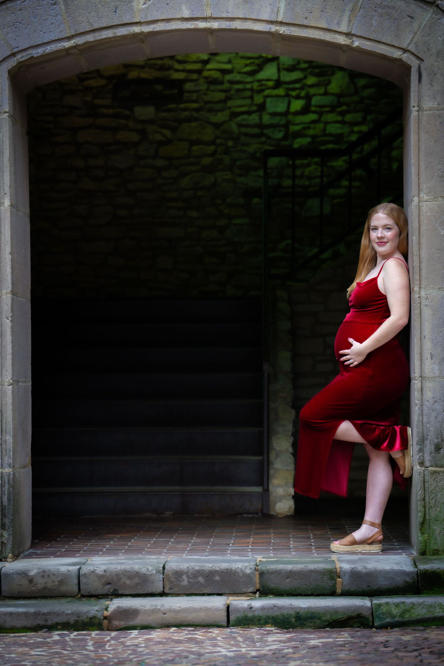 An elegant maternity portrait featuring someone in red velvet standing in a stone archway.