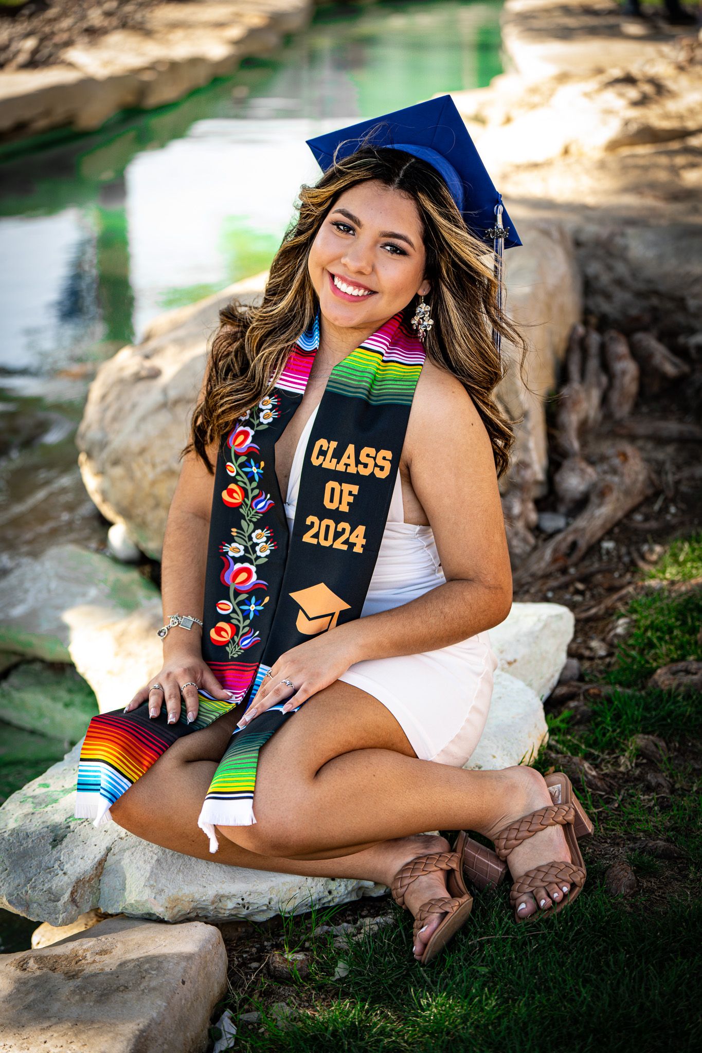 Graduate in blue cap and gown poses on rocks near water with decorated graduation stoles.
