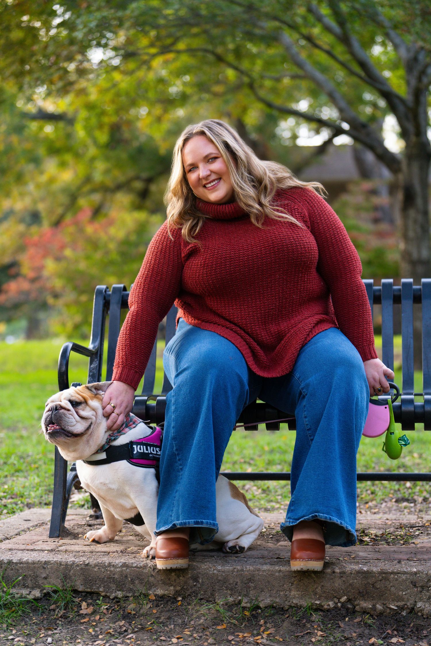 Person in red sweater sits on park bench with English bulldog during autumn.