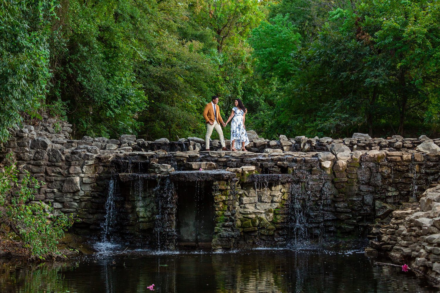 A couple stands atop a scenic stone waterfall structure surrounded by lush green foliage at a natural park.