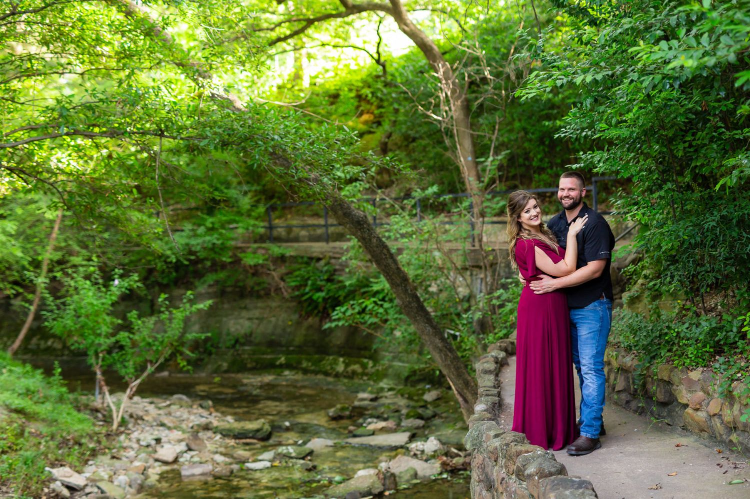 A couple in formal attire embraces on a scenic woodland path beside a rocky stream and lush greenery.