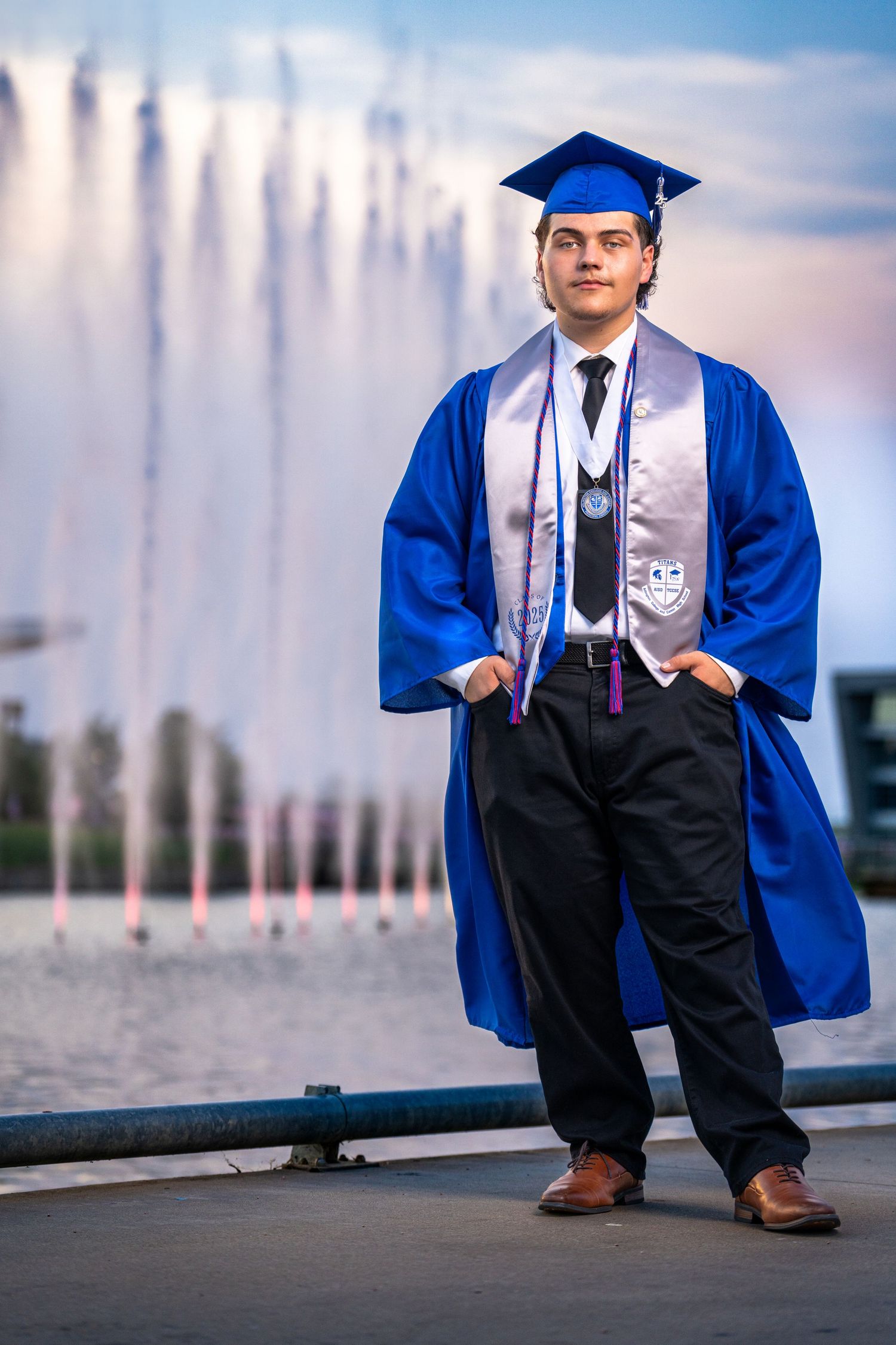Graduate student in blue cap and gown poses confidently in front of a fountain on their graduation day.