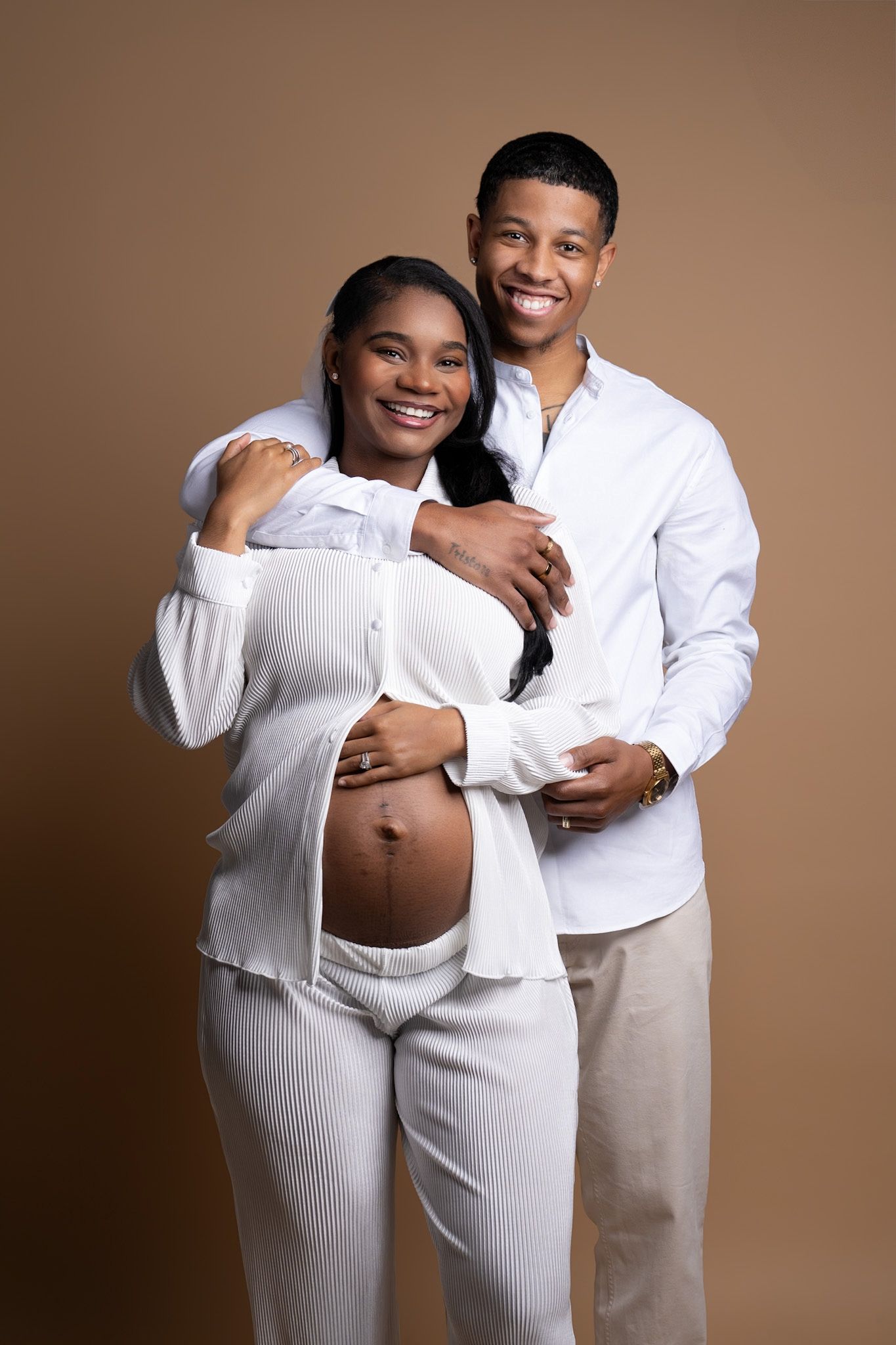 An expecting couple in white outfits poses together against a brown background.