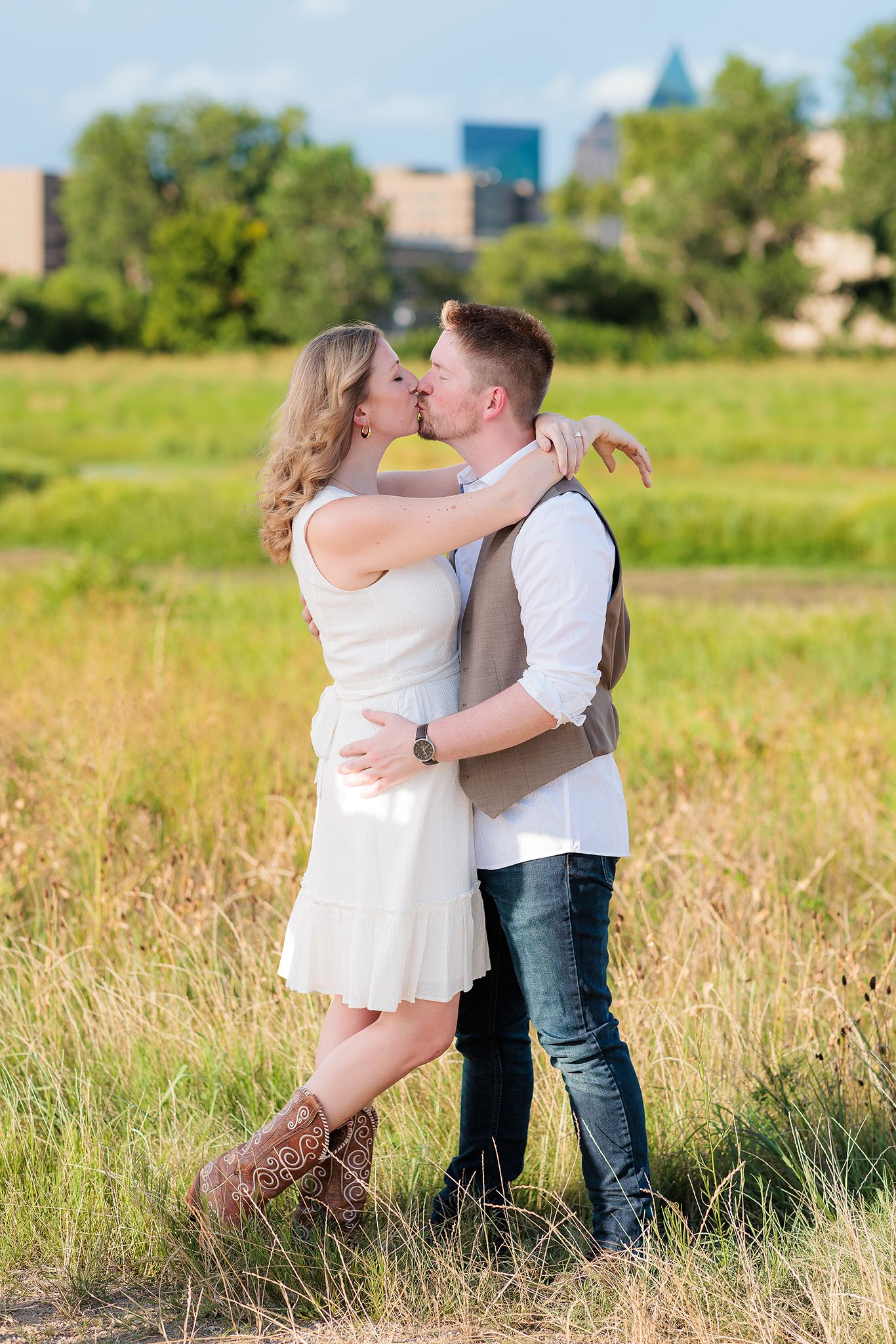 A couple embraces in a sun-lit meadow, she wearing a white dress and boots, he in a vest and jeans.