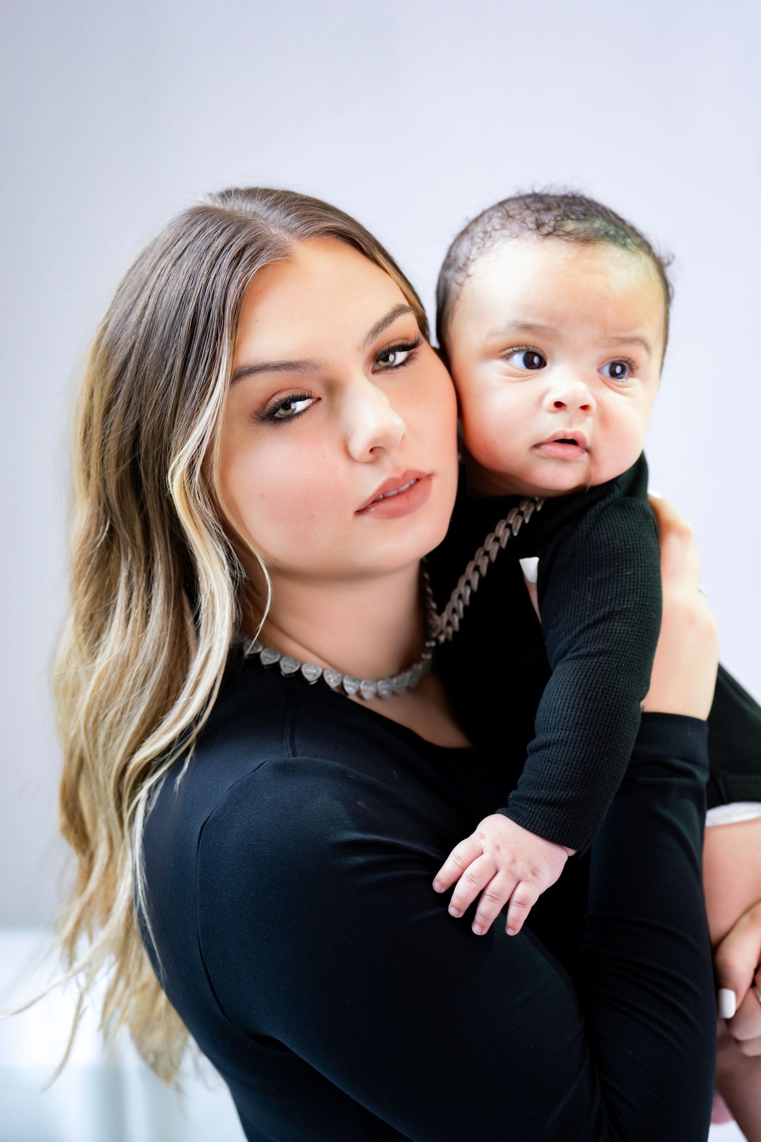 Studio portrait against light background shows woman in black long sleeve top holding small infant in matching black outfit.