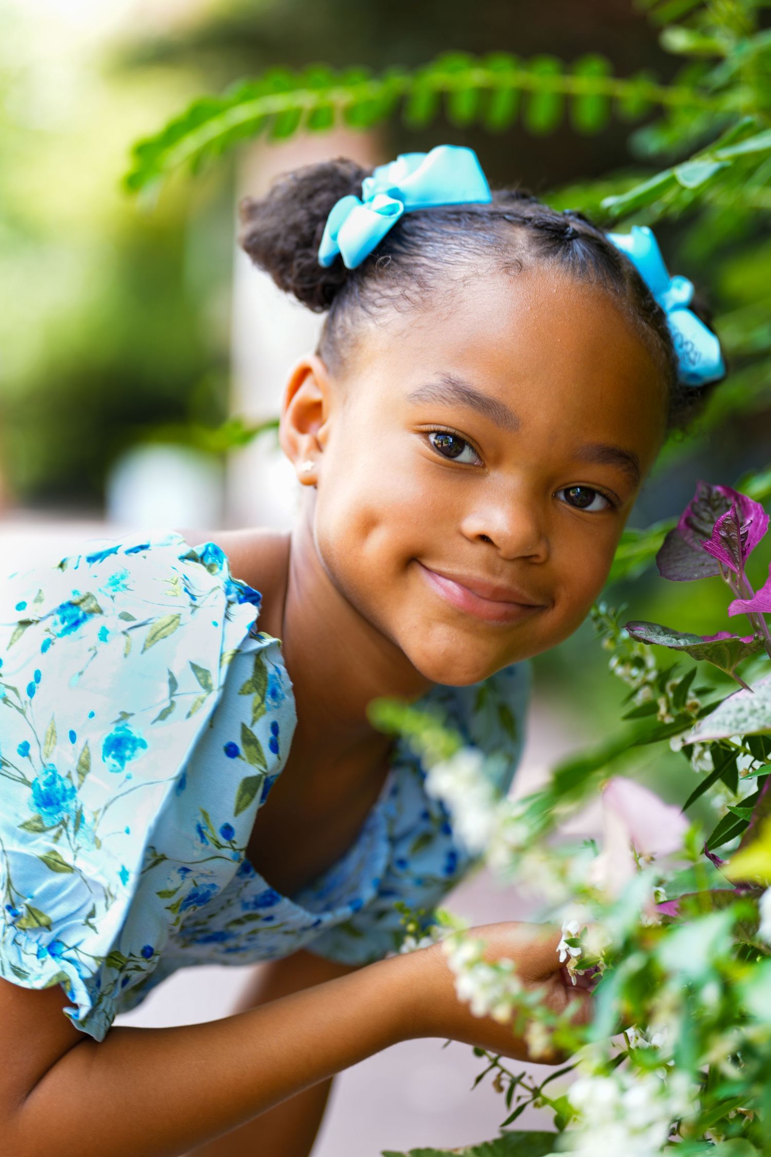 Young model in blue floral dress poses outdoors among greenery with matching blue hair bows.