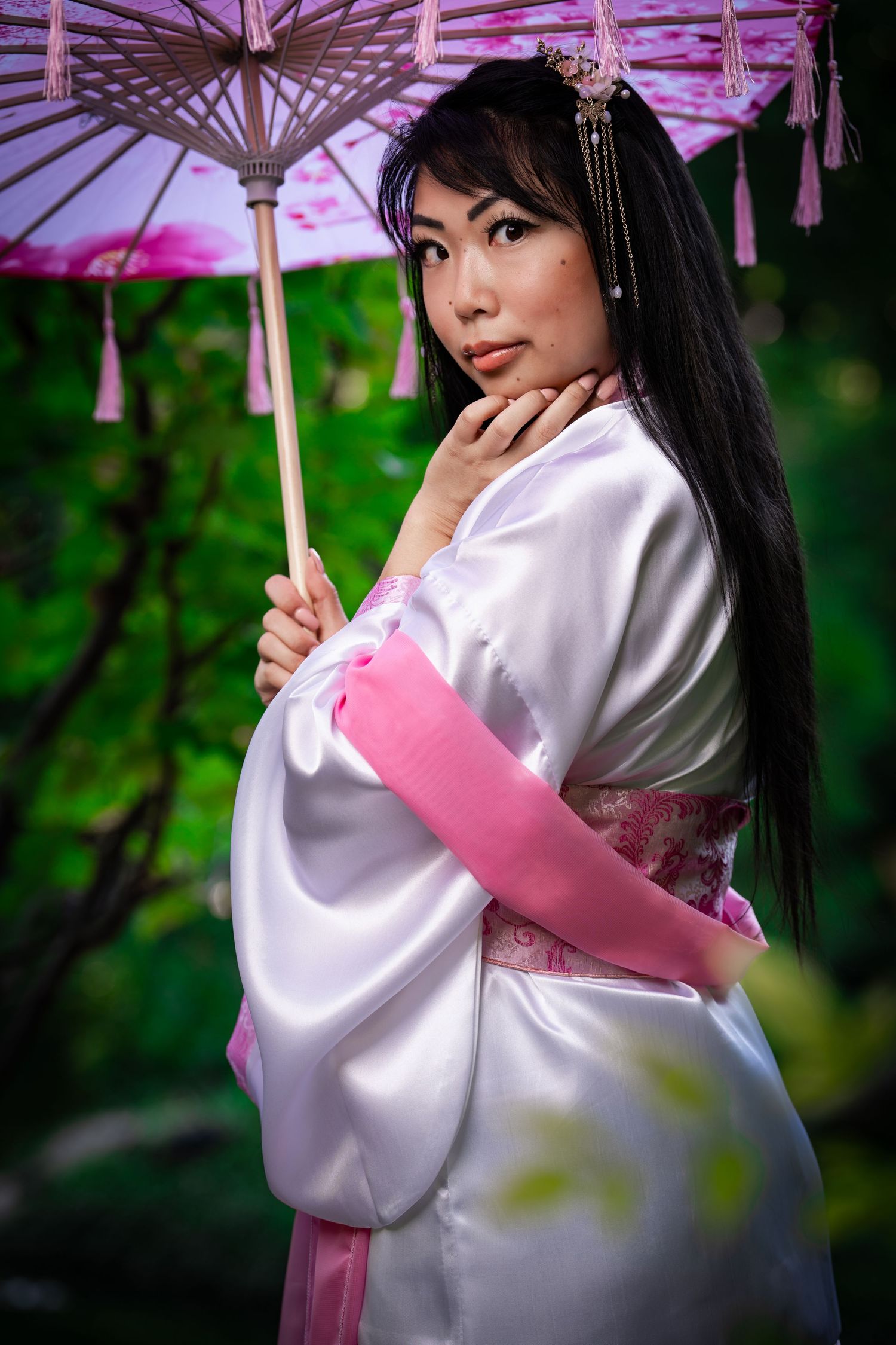 A model in traditional Asian attire poses elegantly with a decorative parasol against a nature backdrop.