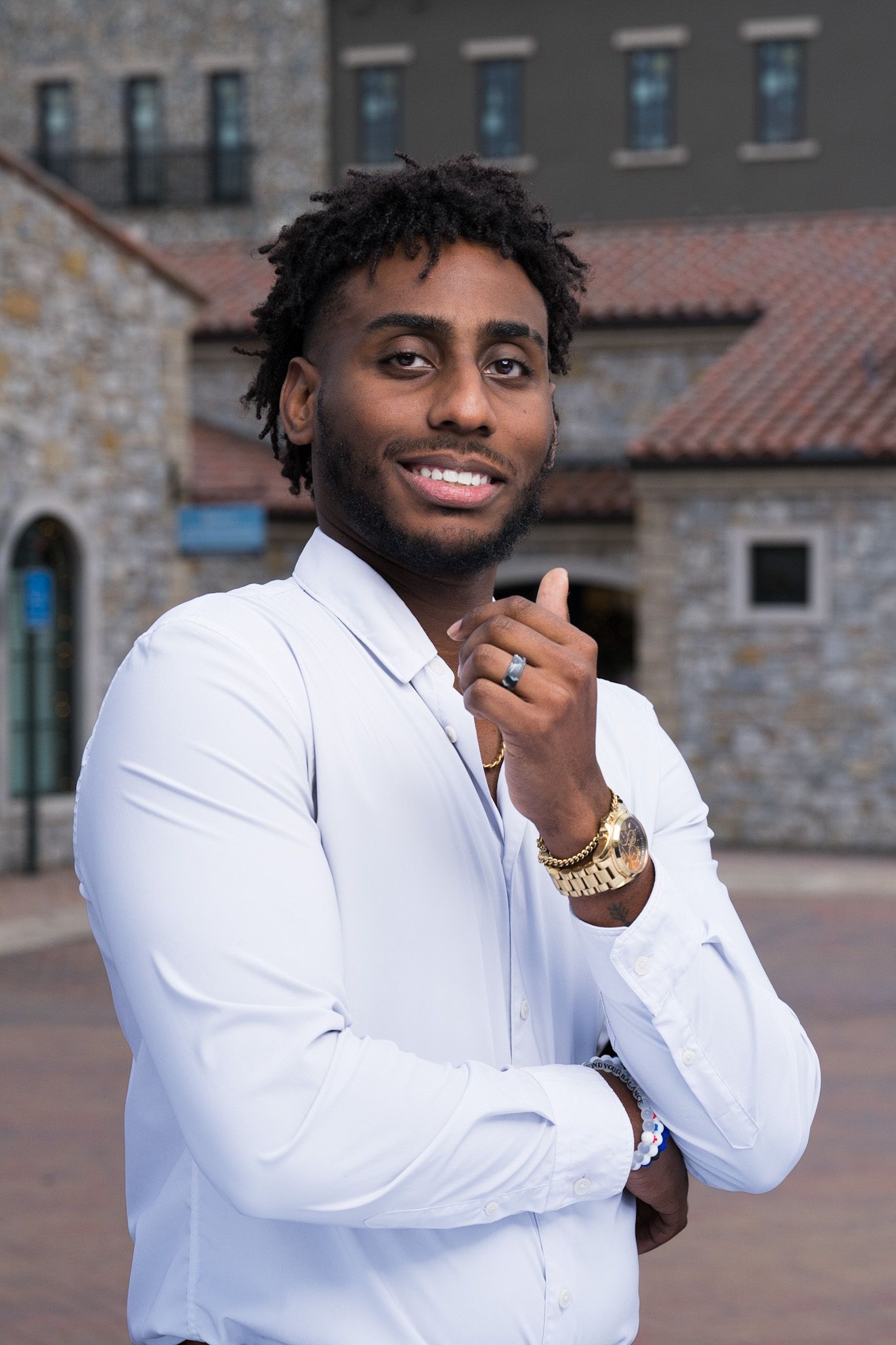 Professional portrait of someone in a crisp white dress shirt with gold jewelry against a stone building backdrop.