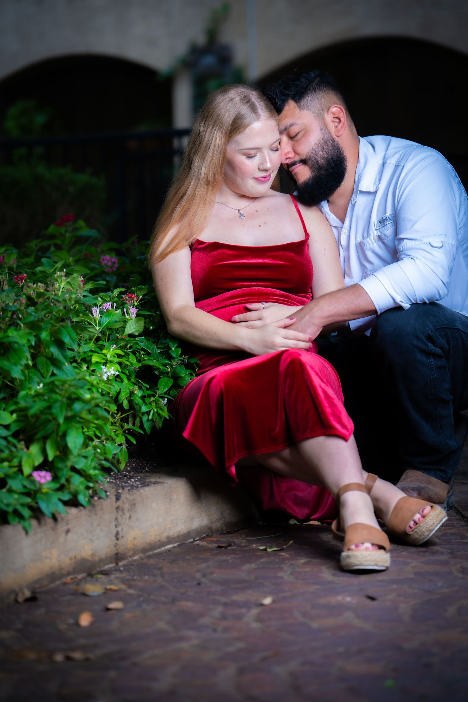A romantic couple shares an intimate moment outdoors, with the woman wearing a red velvet dress.