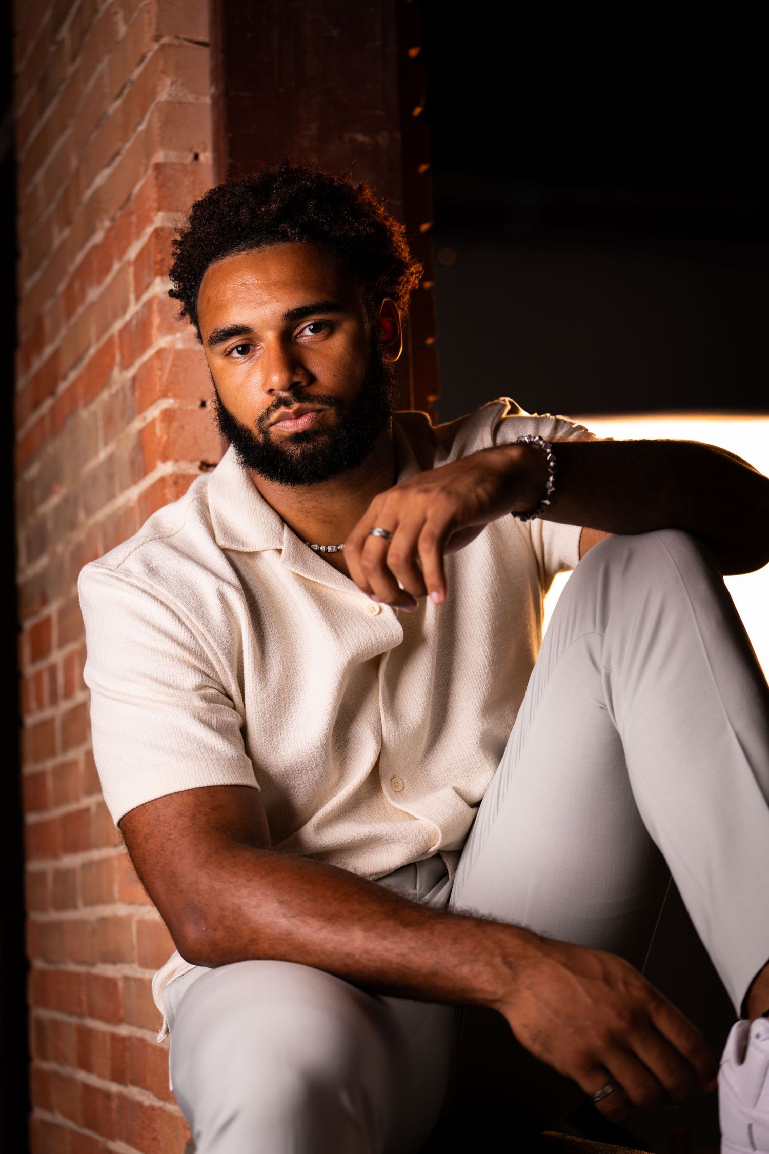 Moody indoor portrait against exposed brick wall in all-white casual attire.