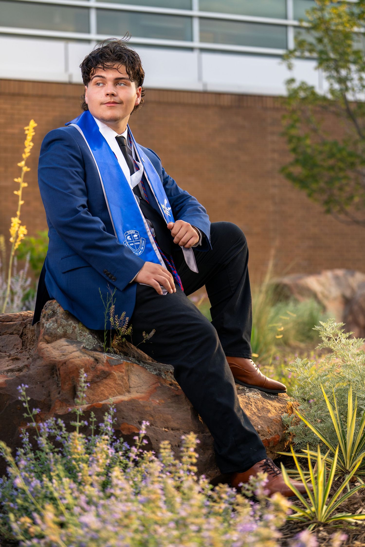 A person in a blue blazer sits casually on a natural wood log surrounded by colorful garden flowers and landscaping.