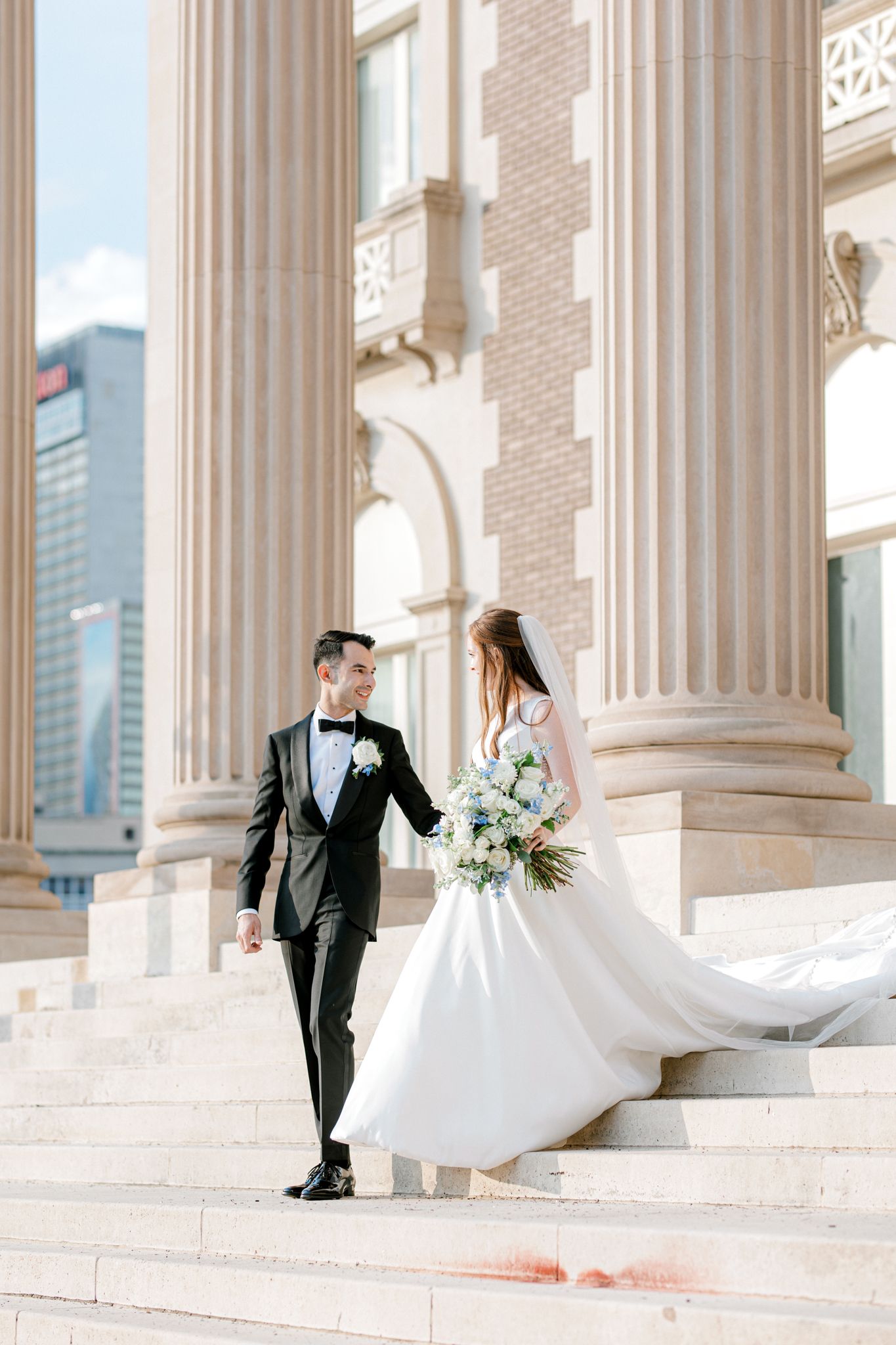 Elegant wedding couple twirls and poses along a grand colonnade with white pillars and modern city backdrop.