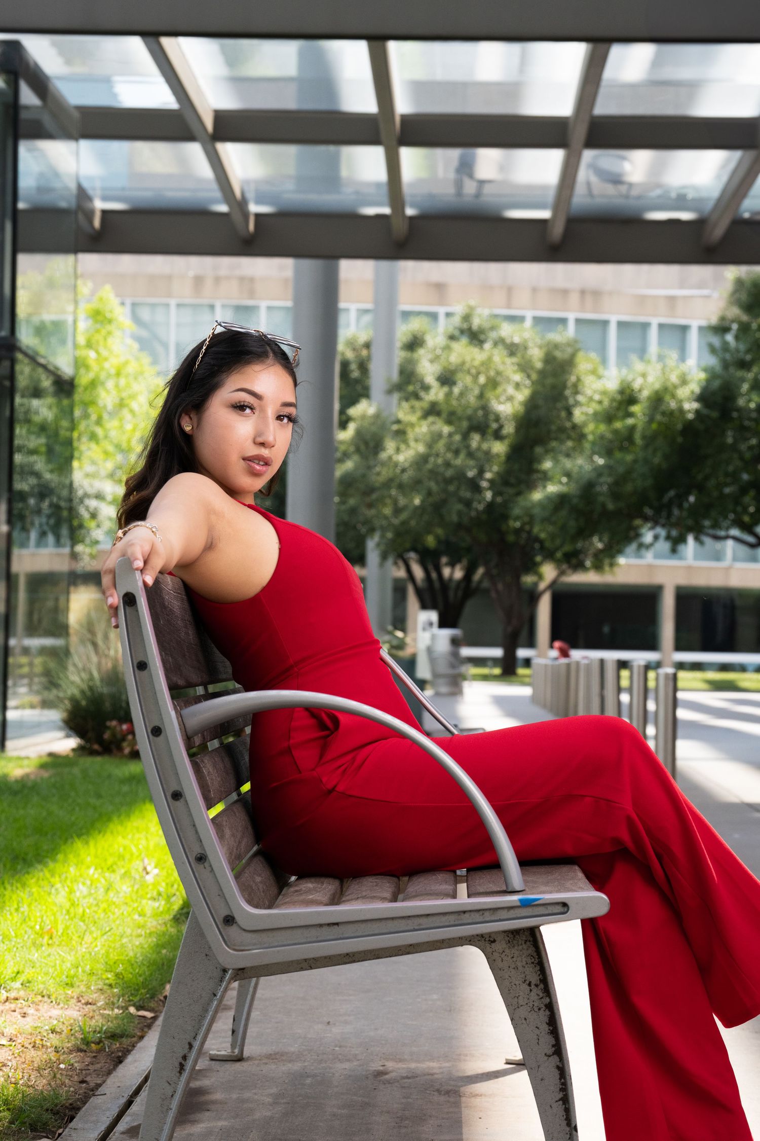 Model in a flowing red dress poses elegantly on a modern bench in an outdoor setting with greenery in the background.