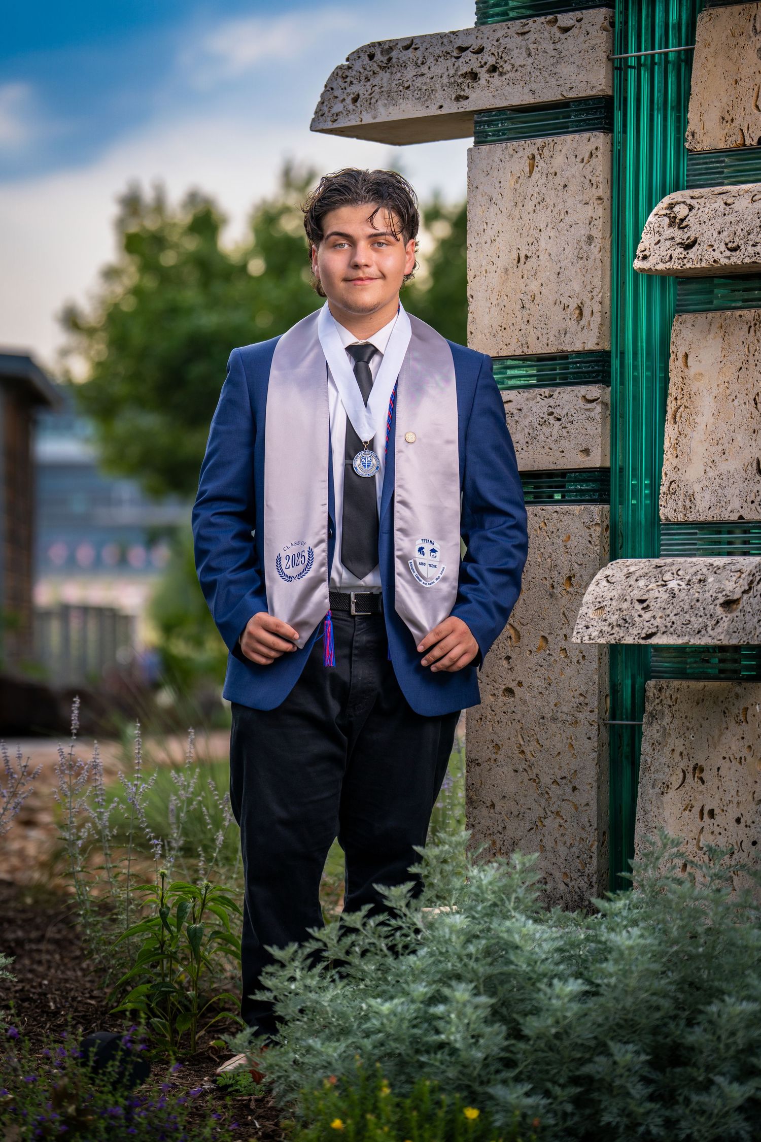 A graduating student in a navy suit and academic stole stands beside a stone pillar and green gate in an outdoor setting.