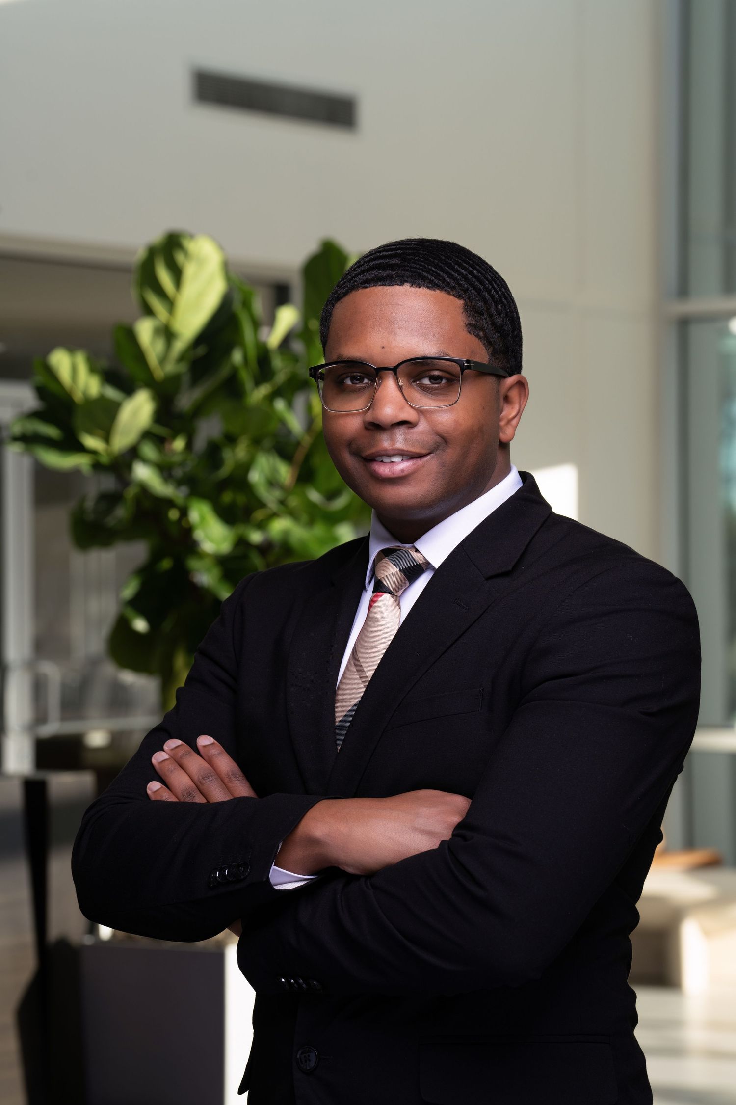 Professional headshot of person in business suit with arms crossed in office lobby.