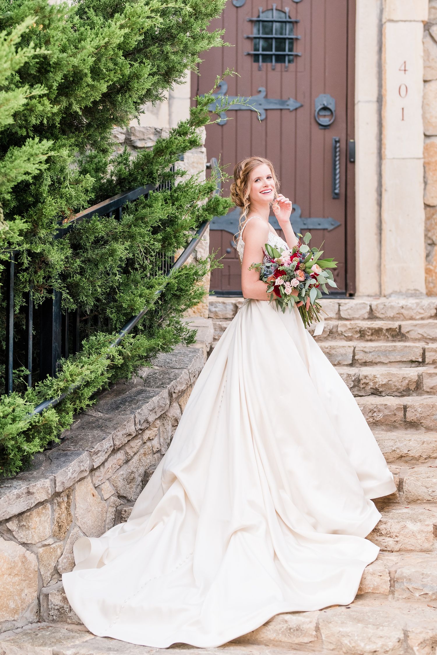 A bride in a flowing white ballgown stands by rustic stone steps and wooden doors holding a colorful bouquet.