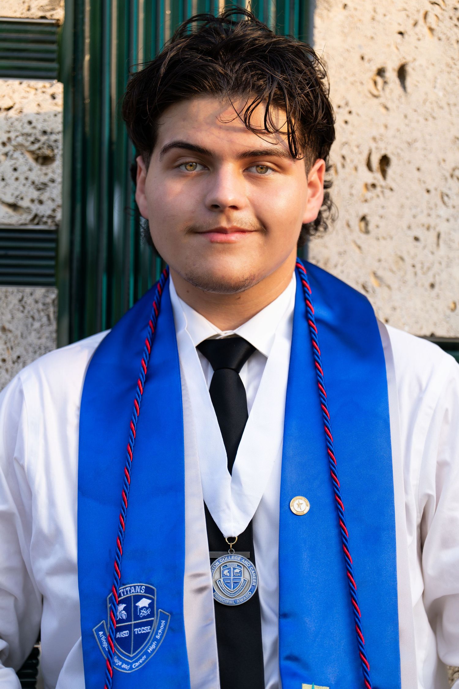 High school graduate wearing a white dress shirt, black tie, blue graduation stole with red cords against a stone wall.