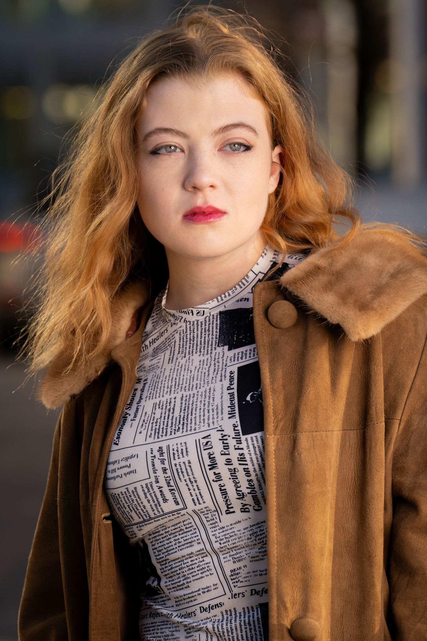 Model wearing a newspaper print top and brown fur-trimmed coat poses for a portrait in natural light.