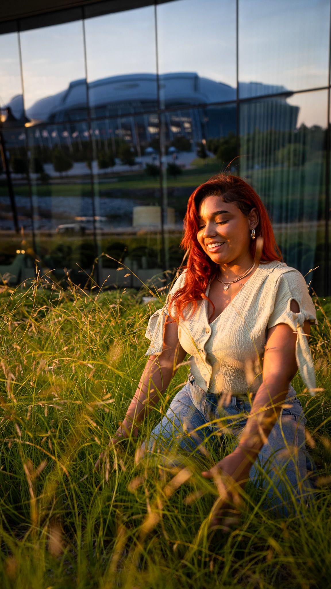 Person with vibrant red hair sitting in tall grass at sunset with building in background.