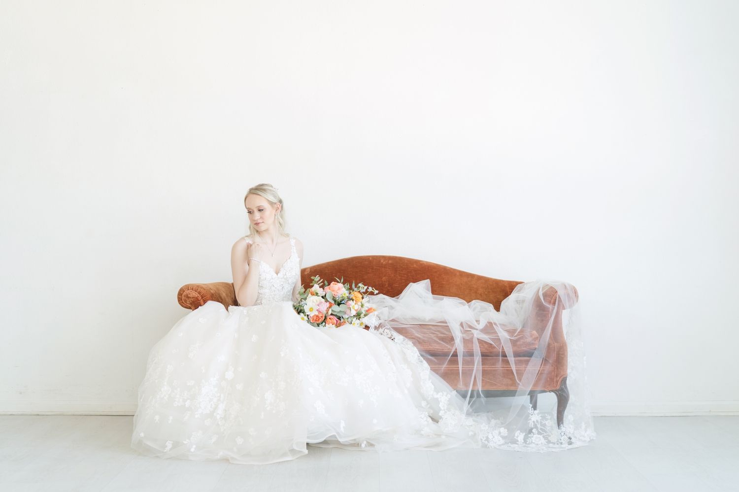 A delicate white wedding dress with lace details drapes elegantly across a vintage brown settee in a bright studio setting.