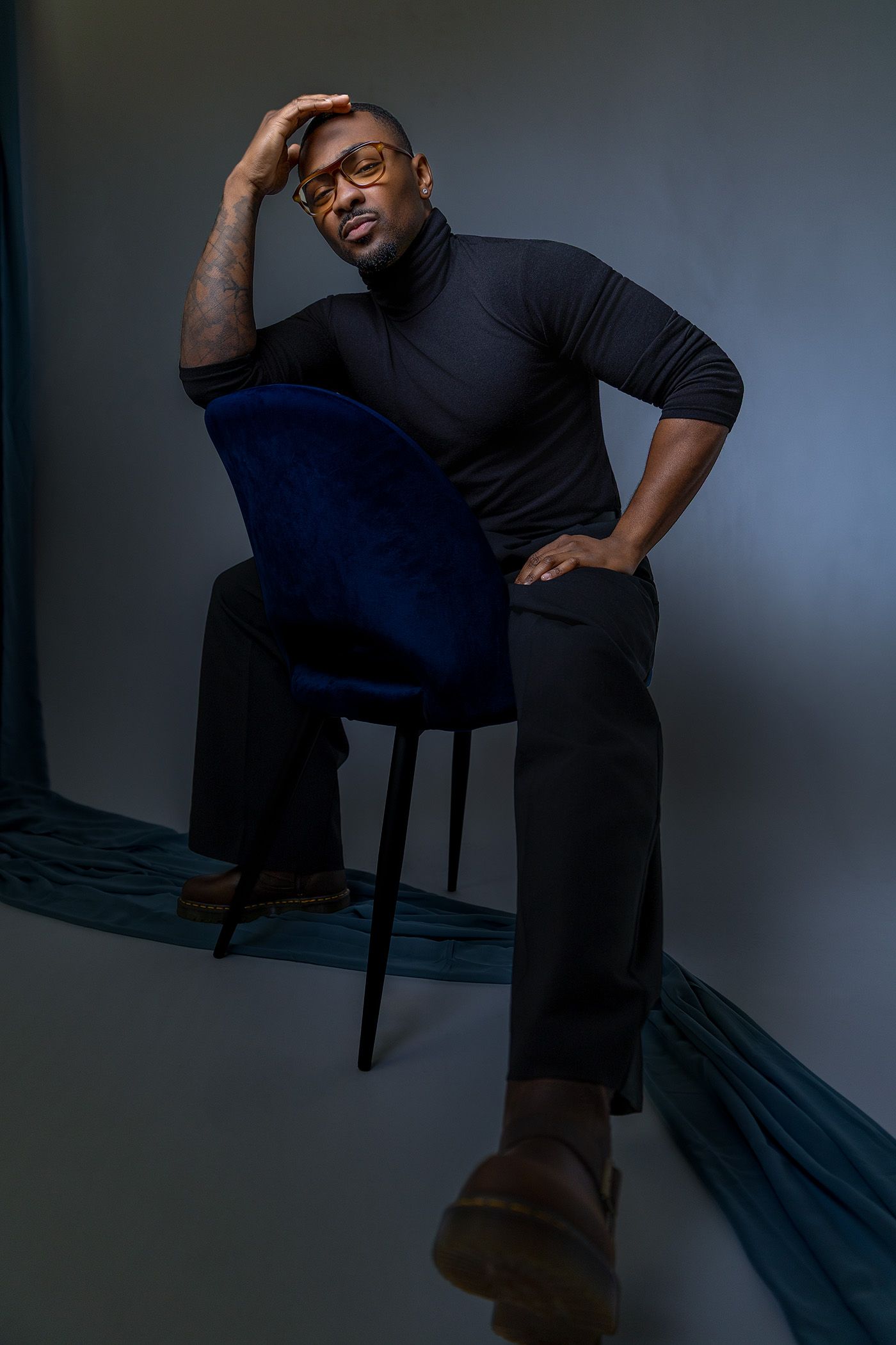 Man in black turtleneck and glasses sitting on blue velvet chair in studio portrait.