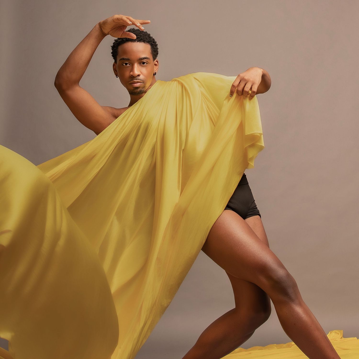 Dancer in black shorts gracefully poses with flowing yellow fabric against neutral backdrop.