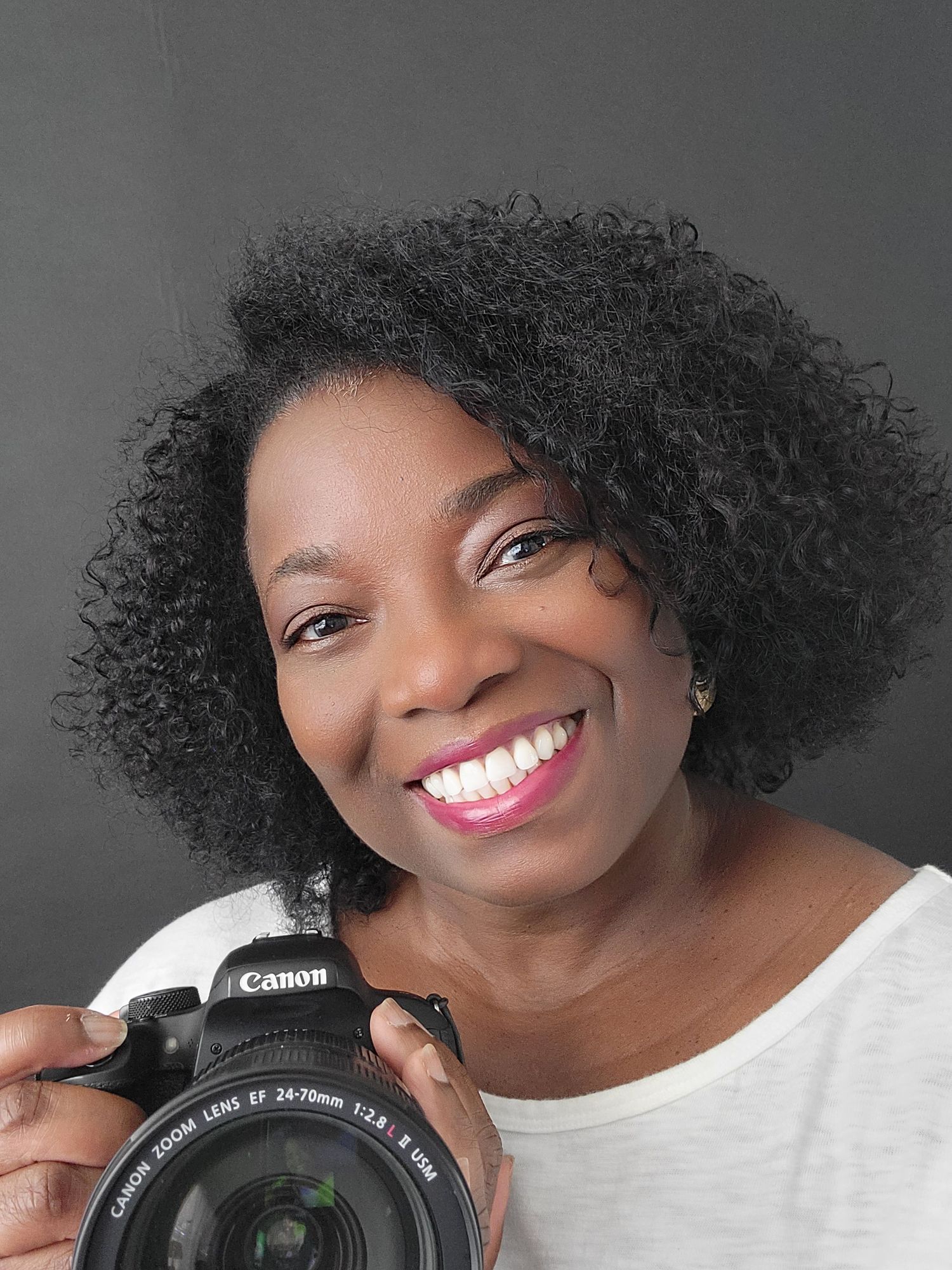 Female Atlanta portrait Photographer in a white top smiles while holding a Canon camera against a gray background.