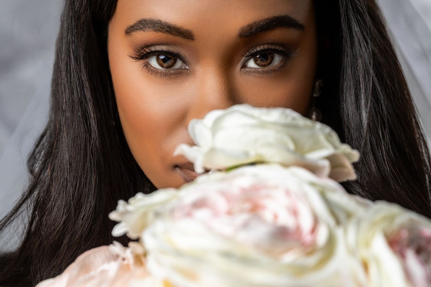 A Black bride holds white and blush peonies near her face, showcasing elegant makeup and a flowing veil.