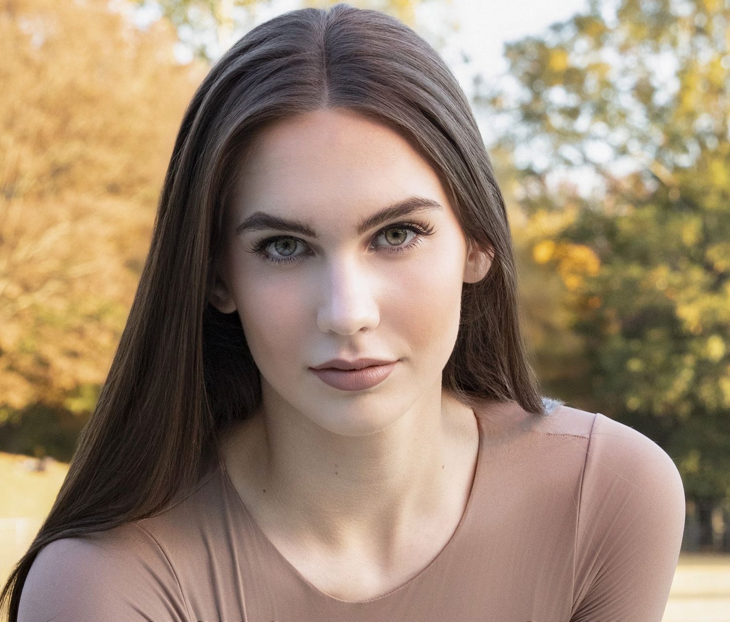 Senior with long brown hair and dark lipstick poses outdoors with autumn trees in the background.