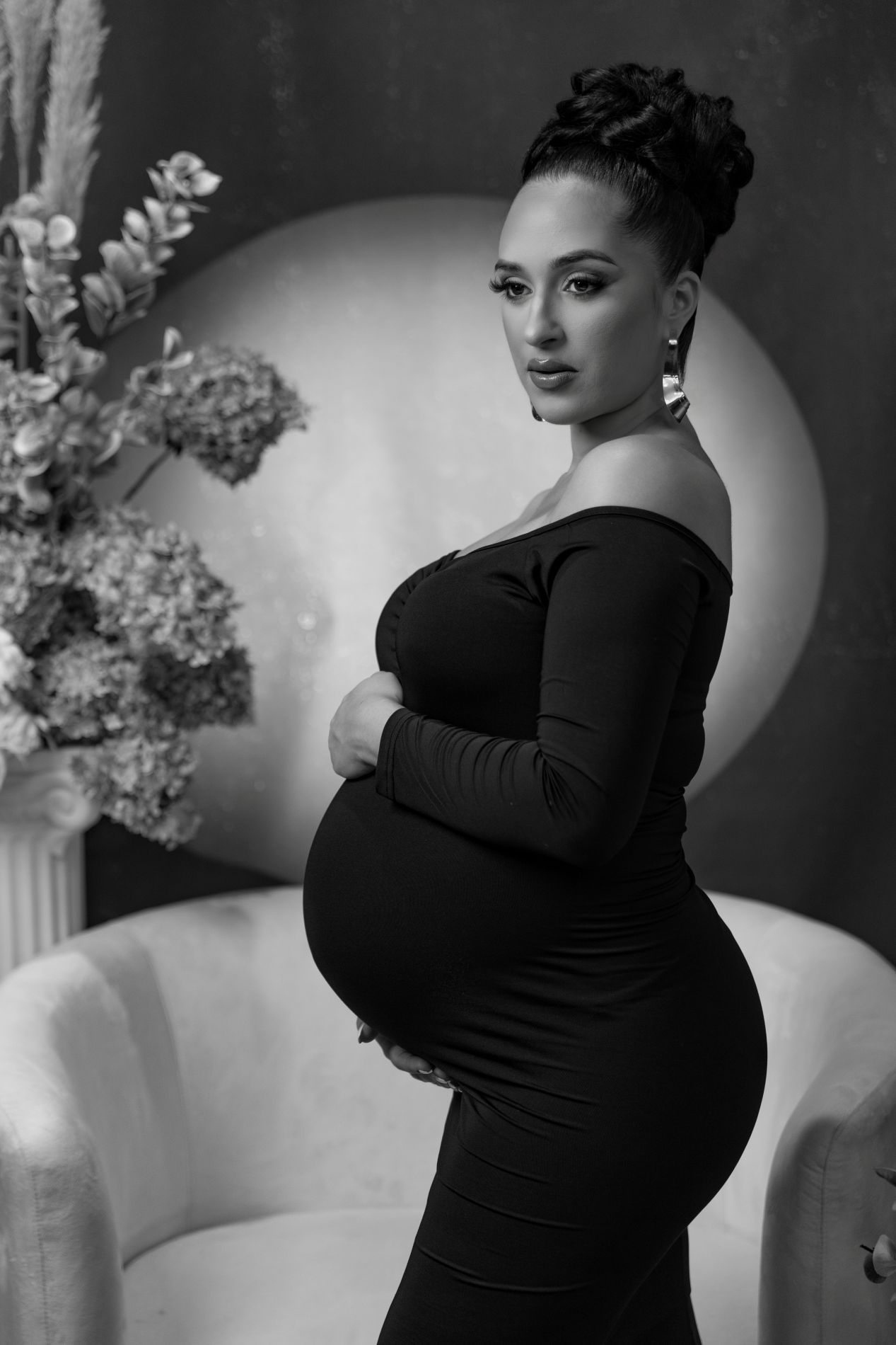 A black and white maternity photo showing a figure in a black dress posing against a light backdrop with flowers.
