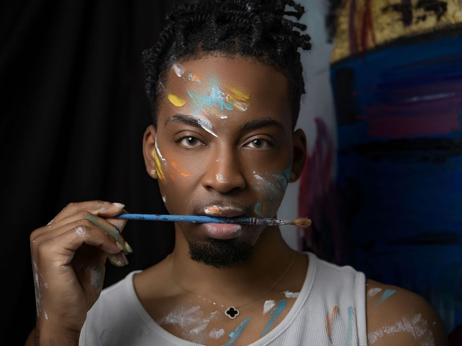 Young Black male artist holding a paintbrush to his lips, face adorned with colorful paint splatters, wearing a white tank top.