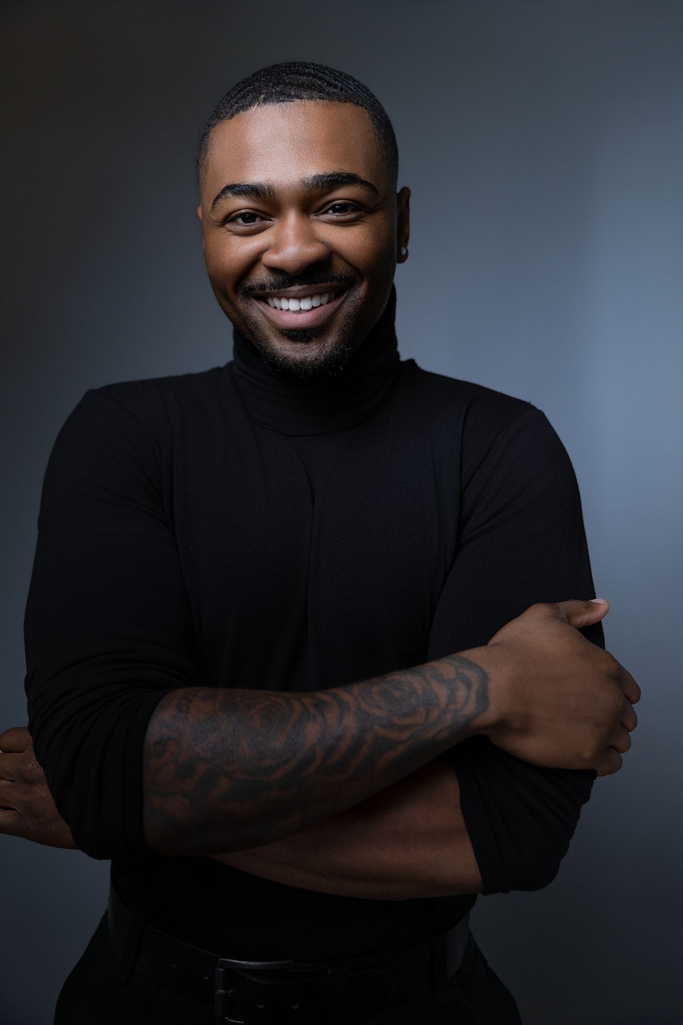 Smiling Black man with tattoos crosses arms, wearing black turtleneck, studio portrait on dark background.