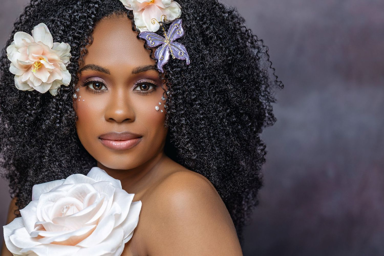 A beautiful Black woman with curly hair adorned with flowers and a butterfly, holding a white rose.