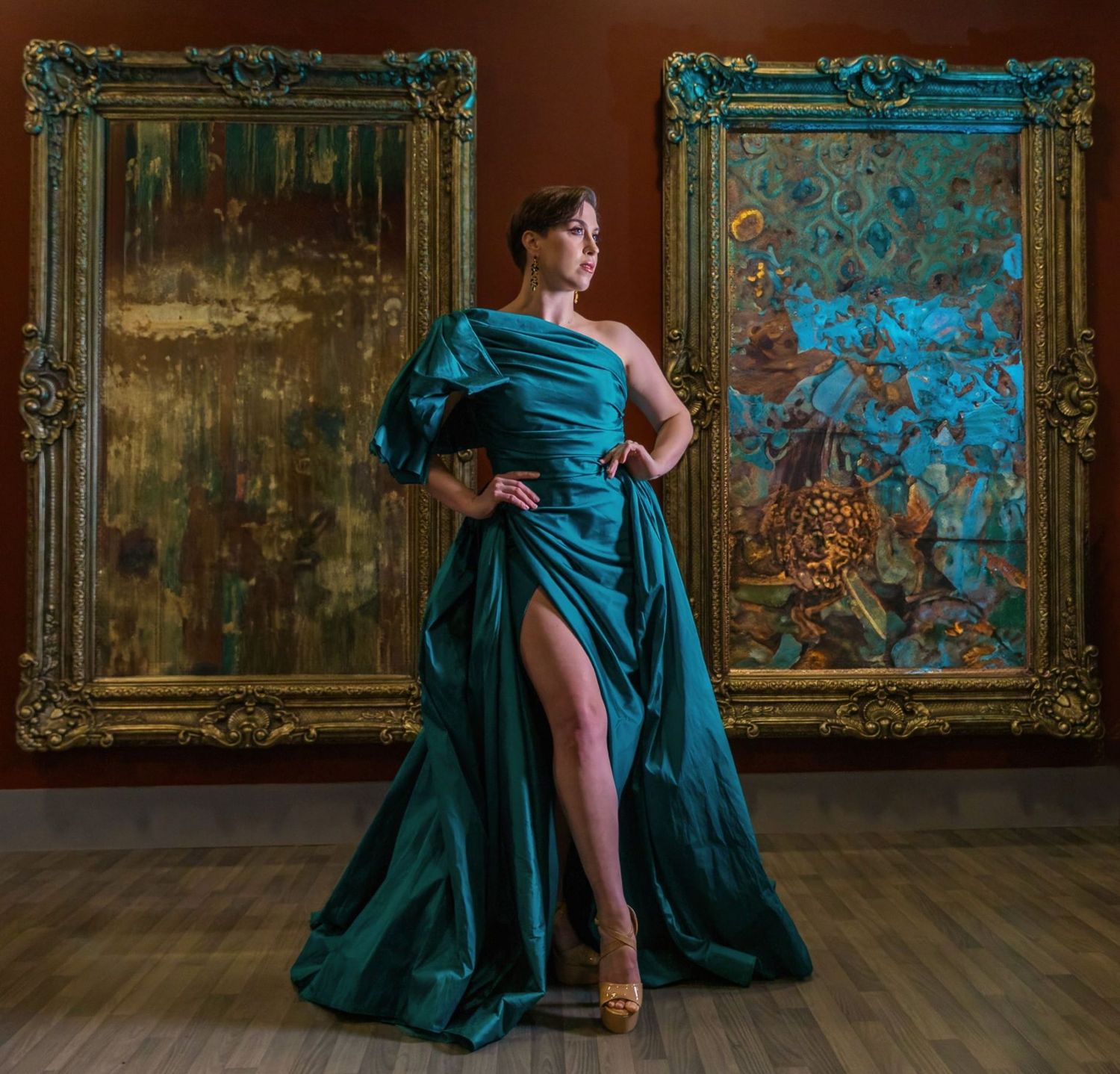 Woman in elegant teal gown poses between ornate gold frames in gallery setting.