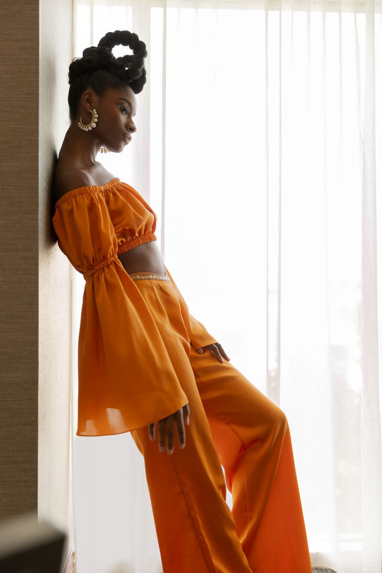 Model wearing vibrant orange off-shoulder ensemble poses in sunlit window setting in Atlanta hotel